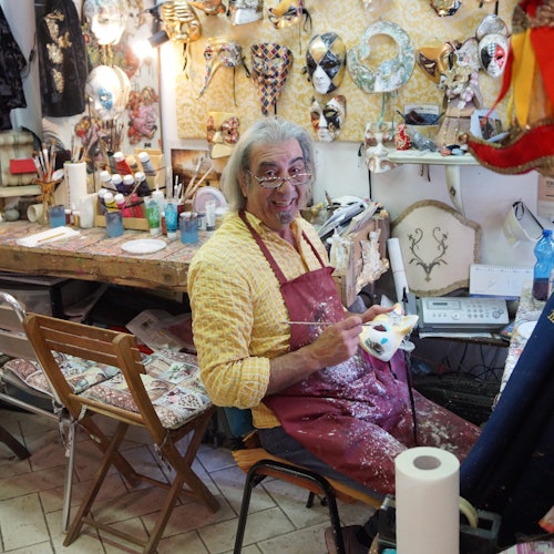 Un uomo sorridente in un laboratorio, con indosso un grembiule rosso, dipinge una maschera. Lo spazio è pieno di materiali artistici e maschere decorate.