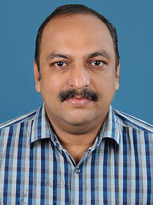 A woman with curly hair and glasses smiles warmly, wearing a peach dress. She's in a sunlit room filled with lush green plants, creating a cozy, serene atmosphere.