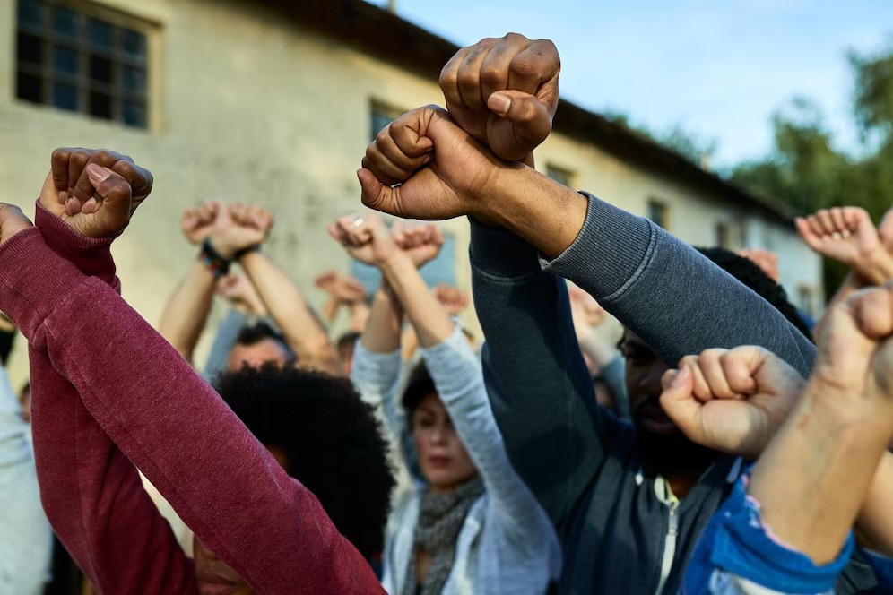 A imagem mostra um grupo de pessoas com os punhos erguidos, um gesto simbólico de resistência e solidariedade. Cada pessoa na foto levanta o braço com o punho fechado, sugerindo uma manifestação ou protesto em grupo. A diversidade das pessoas e a força do gesto destacam um movimento coletivo, possivelmente relacionado à luta por direitos civis ou igualdade racial. O cenário ao fundo é um prédio simples, com a cena focando principalmente nos braços erguidos, enfatizando a unidade e o poder da ação coletiva.