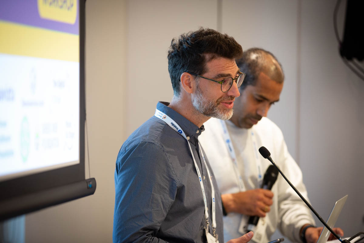 Workshop: Matthieu speaking on stage; Sunnil beside him with a laptop and handheld mic; blurred logo screen on the left.