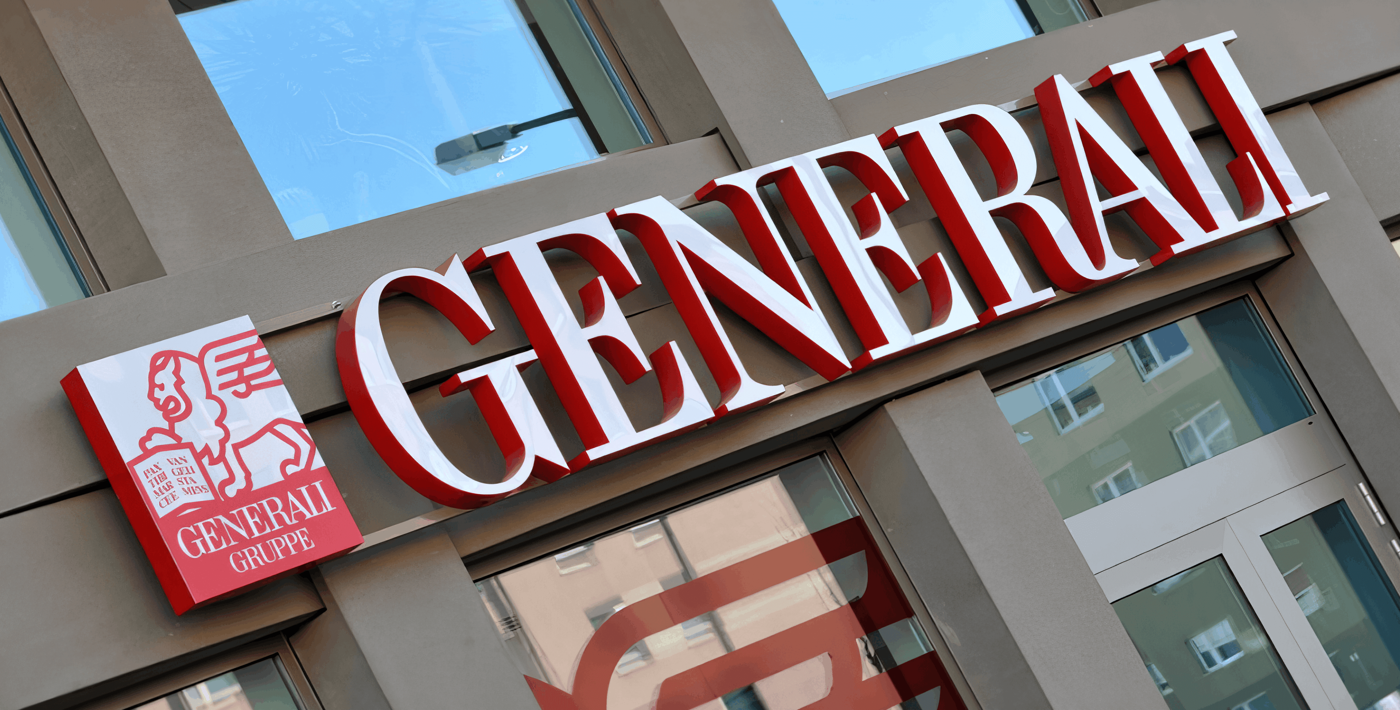 MW Interior - Nahaufnahme des roten Generali-Firmenschilds mit Löwen-Logo an moderner Gebäudefassade" Beschreibung: Markanter Außenauftritt der Generali Gruppe – ein namhafter Referenzkunde, für dessen Büro- und Arbeitsumgebungen hochwertige Einrichtungslösungen realisiert wurden.