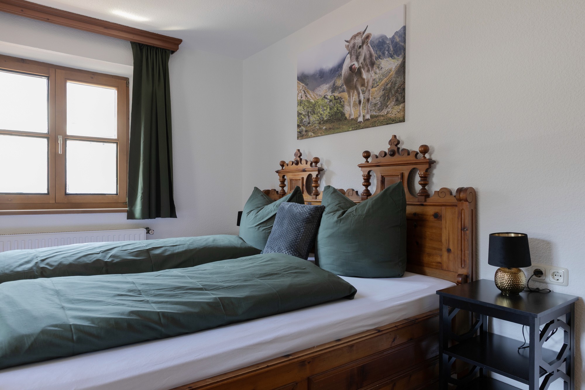 Ein gemütliches Schlafzimmer verfügt über zwei Vintage-Holzbetten mit dunkelgrüner Bettwäsche, einen Nachttisch mit einer schwarzen Lampe und ein Fenster mit einem dunkelgrünen Vorhang, alles ergänzt durch ein Kunstwerk einer Bergziege an der Wand.
