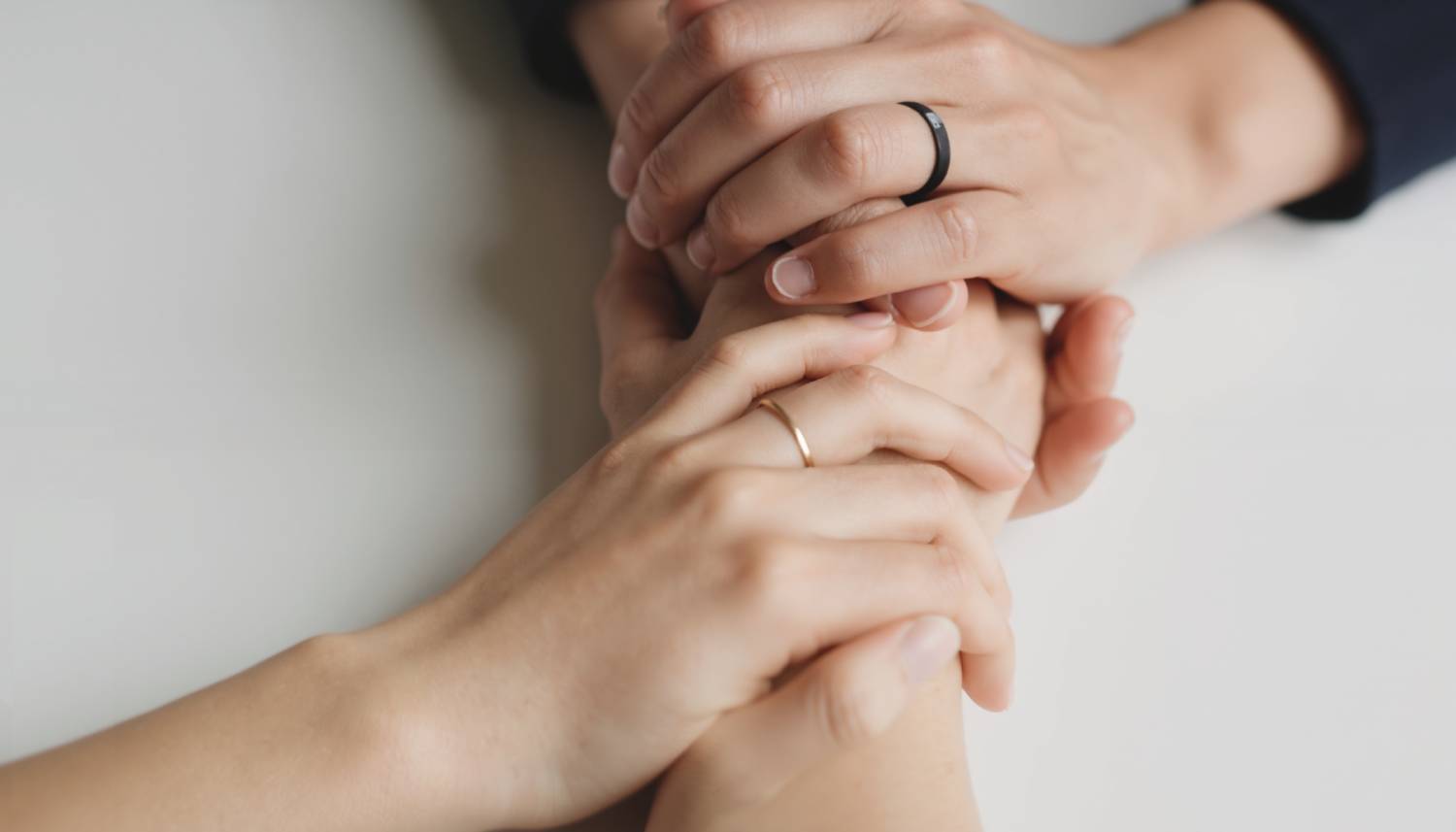 A close-up of two people's hands gently clasped together in a supportive gesture. One person wears a simple gold ring, and the other wears a black band.