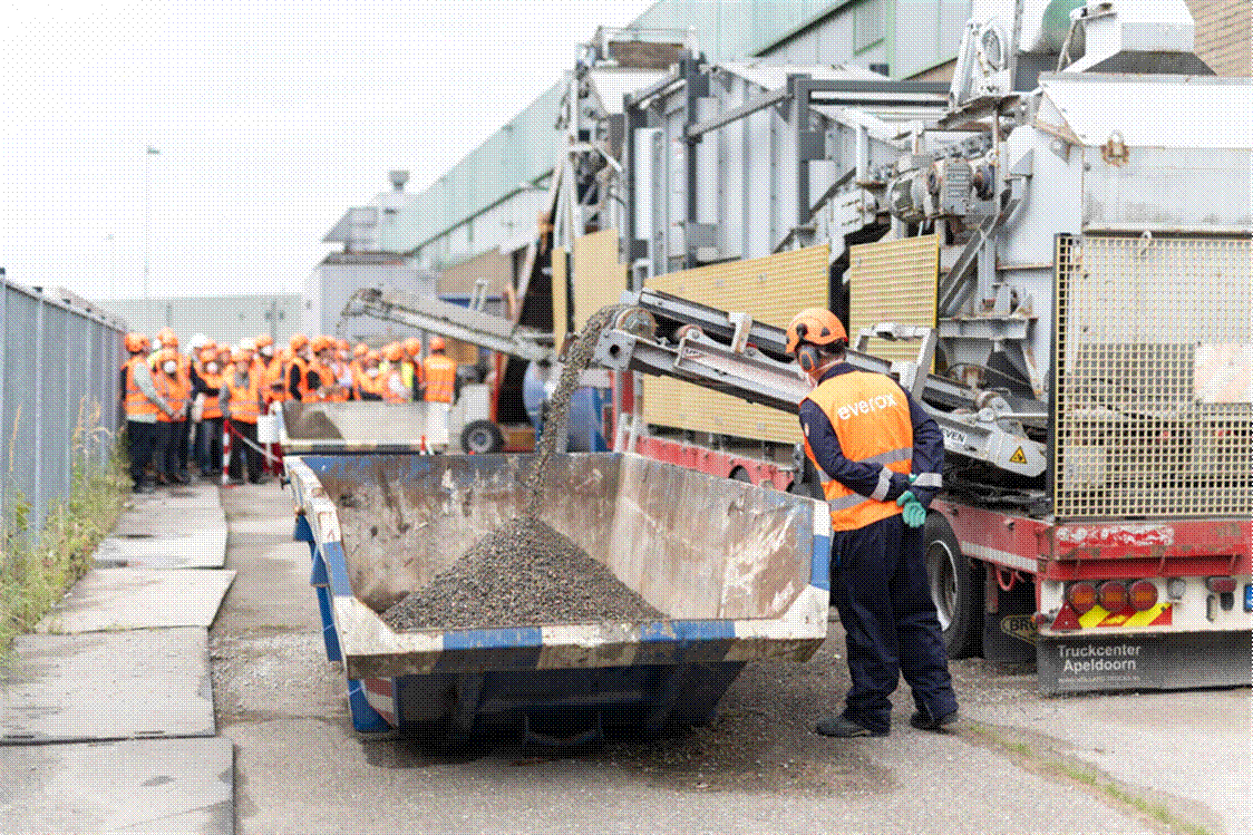 Title: An Everox operator watches recycled aggregate discharge from a mobile processing plant into a skip, - Description: An Everox operator watches recycled aggregate discharge from a mobile processing plant into a skip, with a group of industrial visitors in high-visibility gear looking on.