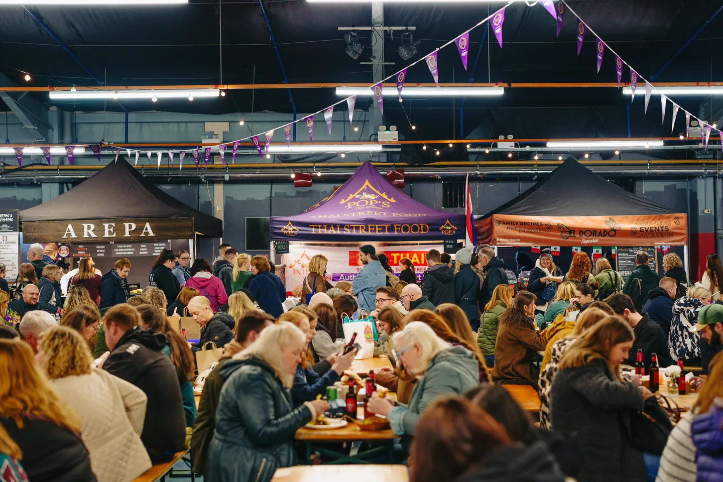 Large food hall scene showing many people sitting and laughing infront of multiple street food exhibitors