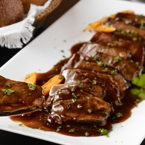 Sliced roast beef covered in a dark gravy, garnished with herbs, on a white rectangular plate. A basket of bread is in the background.