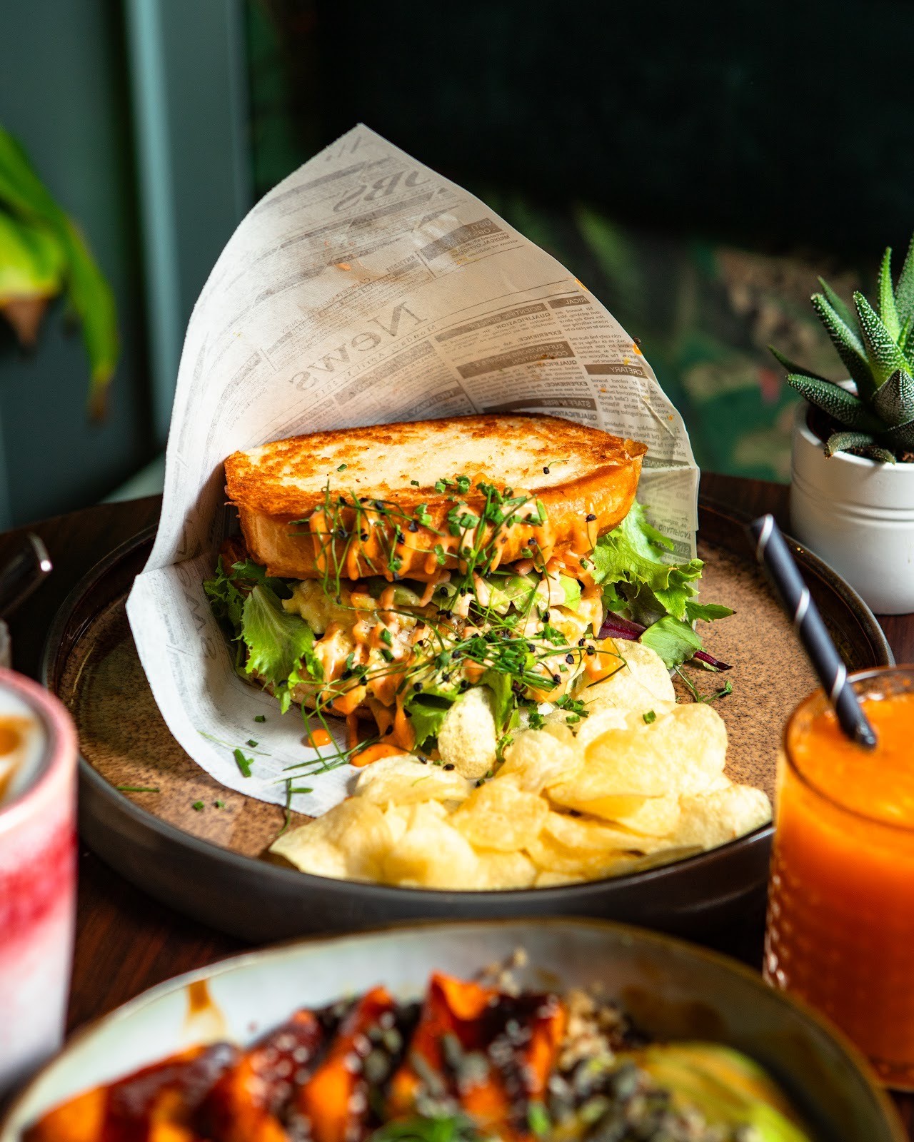 A sandwich wrapped in paper, served with chips and a drink, on a wooden table surrounded by greenery.