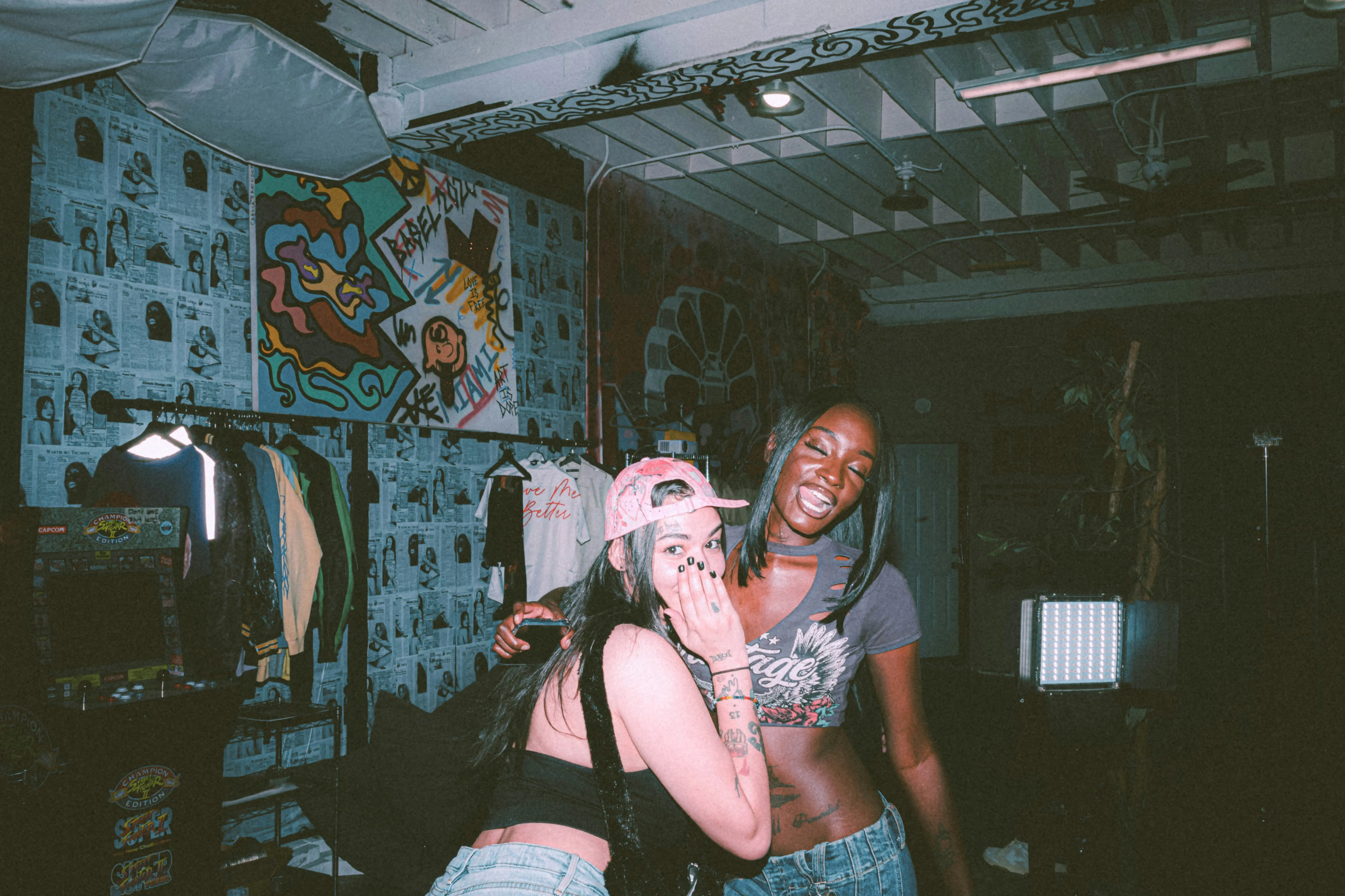 Two friends laughing and posing together in a studio space decorated with graffiti-style wall art, a clothing rack, and ambient lighting.