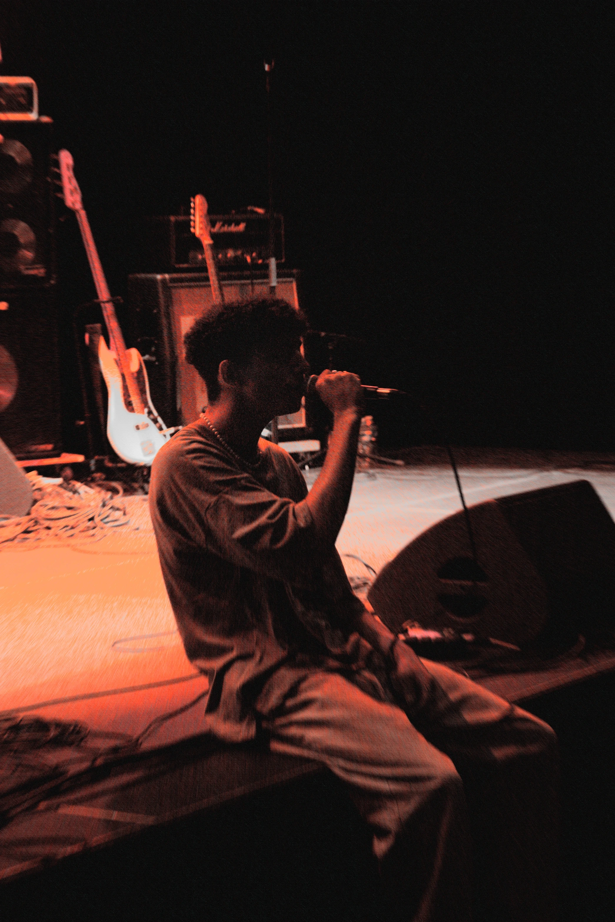 A silhouetted performer sits on stage, passionately singing into a microphone against a vibrant orange background.