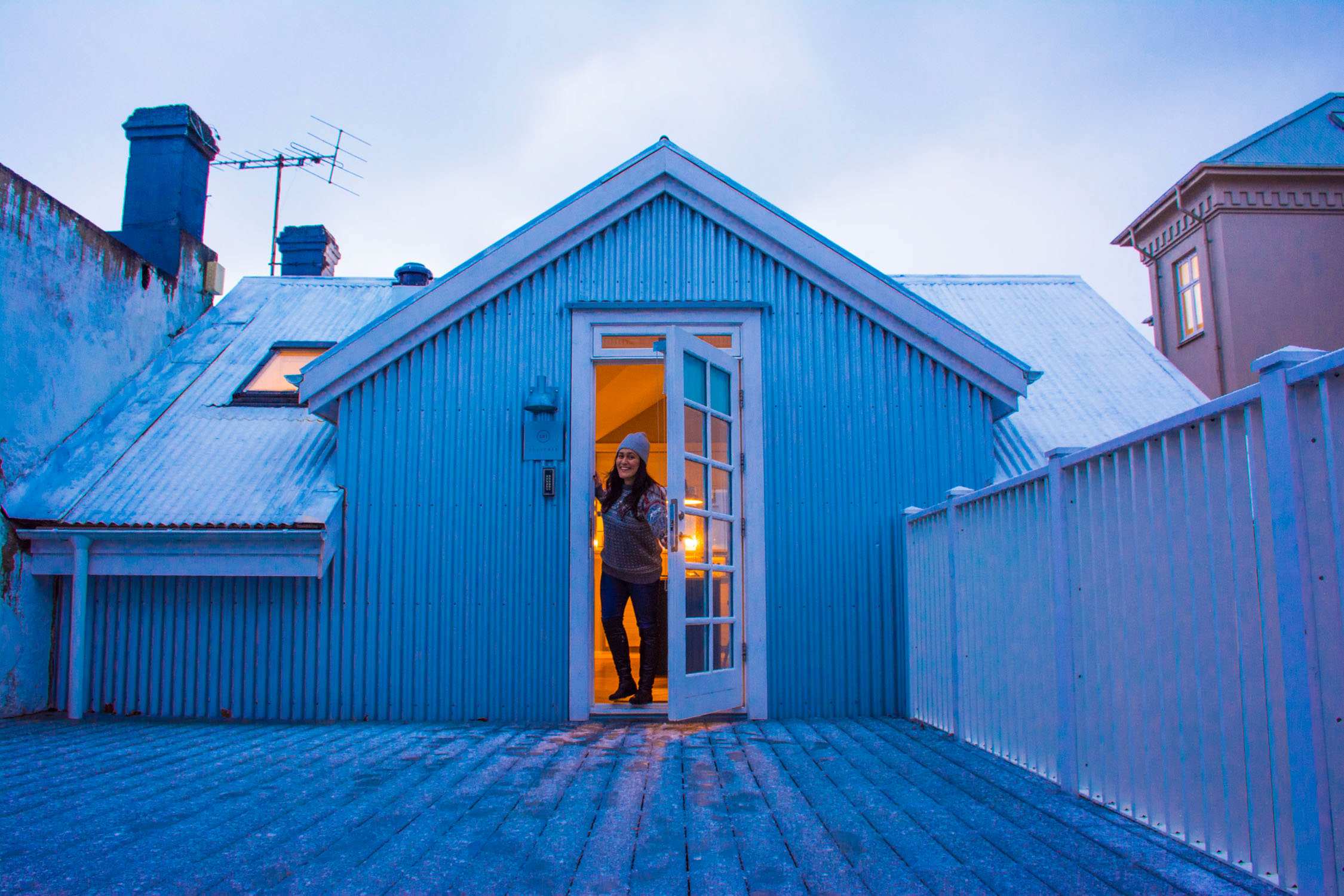 A person in a dark coat stands in an open doorway of a blue wooden cabin with a metal roof under a pale sky.