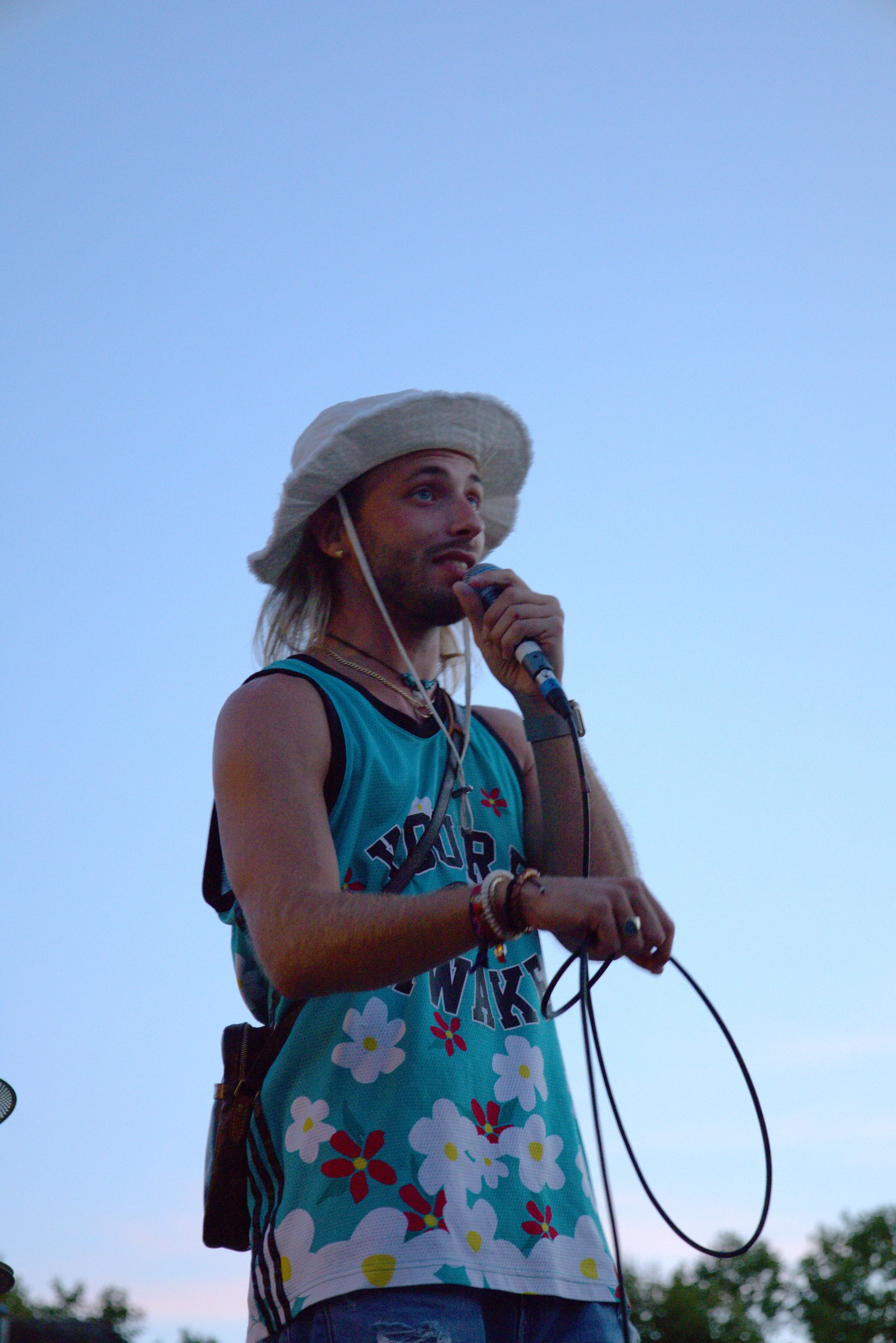 A person wearing a colorful tank top and wide-brimmed hat stands outdoors, speaking into a microphone.