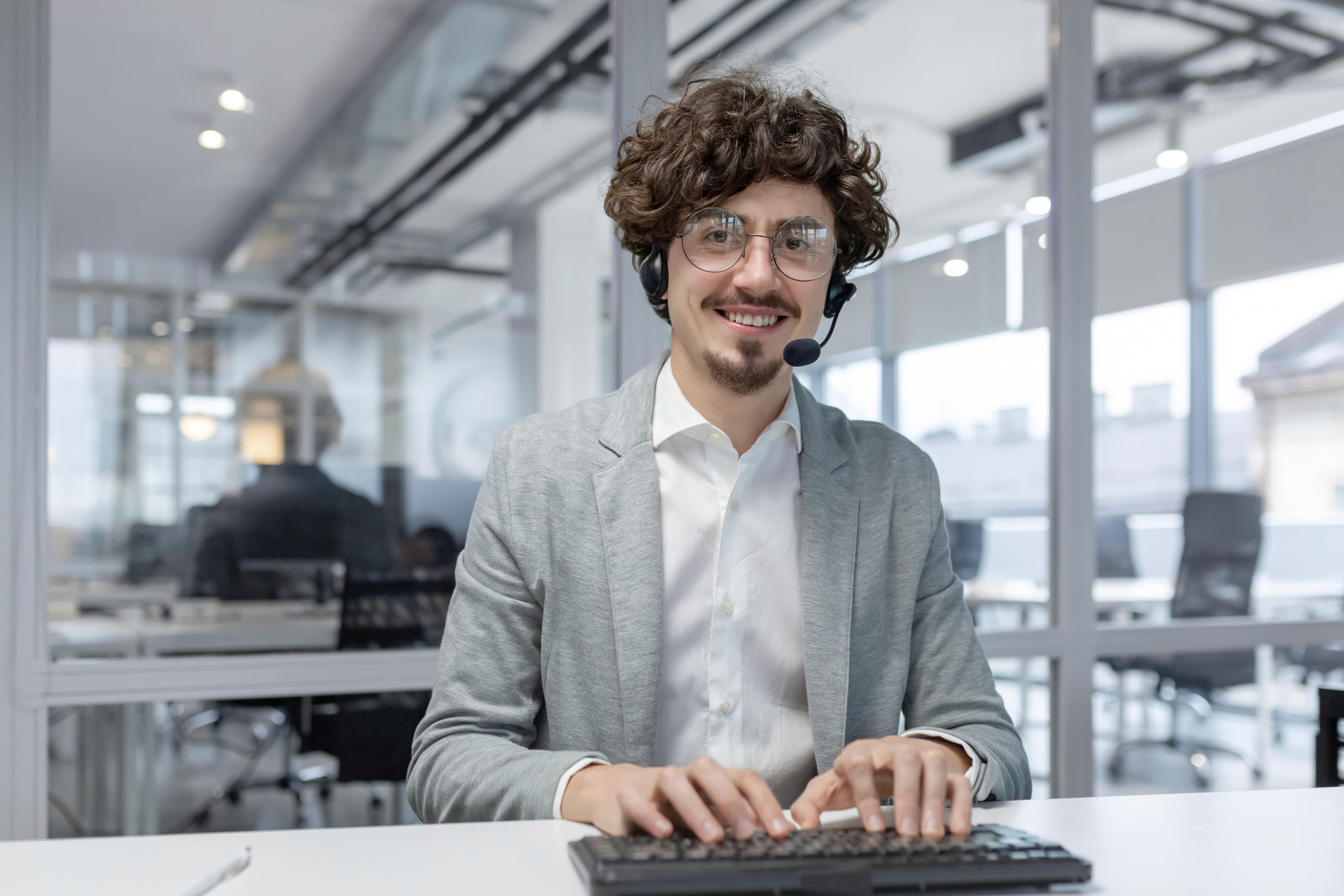 Ein junger, lächelnder Mann mit lockigen Haaren, Brille und einem Headset sitzt an einem weißen Schreibtisch in einem modernen Großraumbüro. Er hat die Hände auf der Tastatur, als ob er gerade tippt, und blickt freundlich direkt in die Kamera.