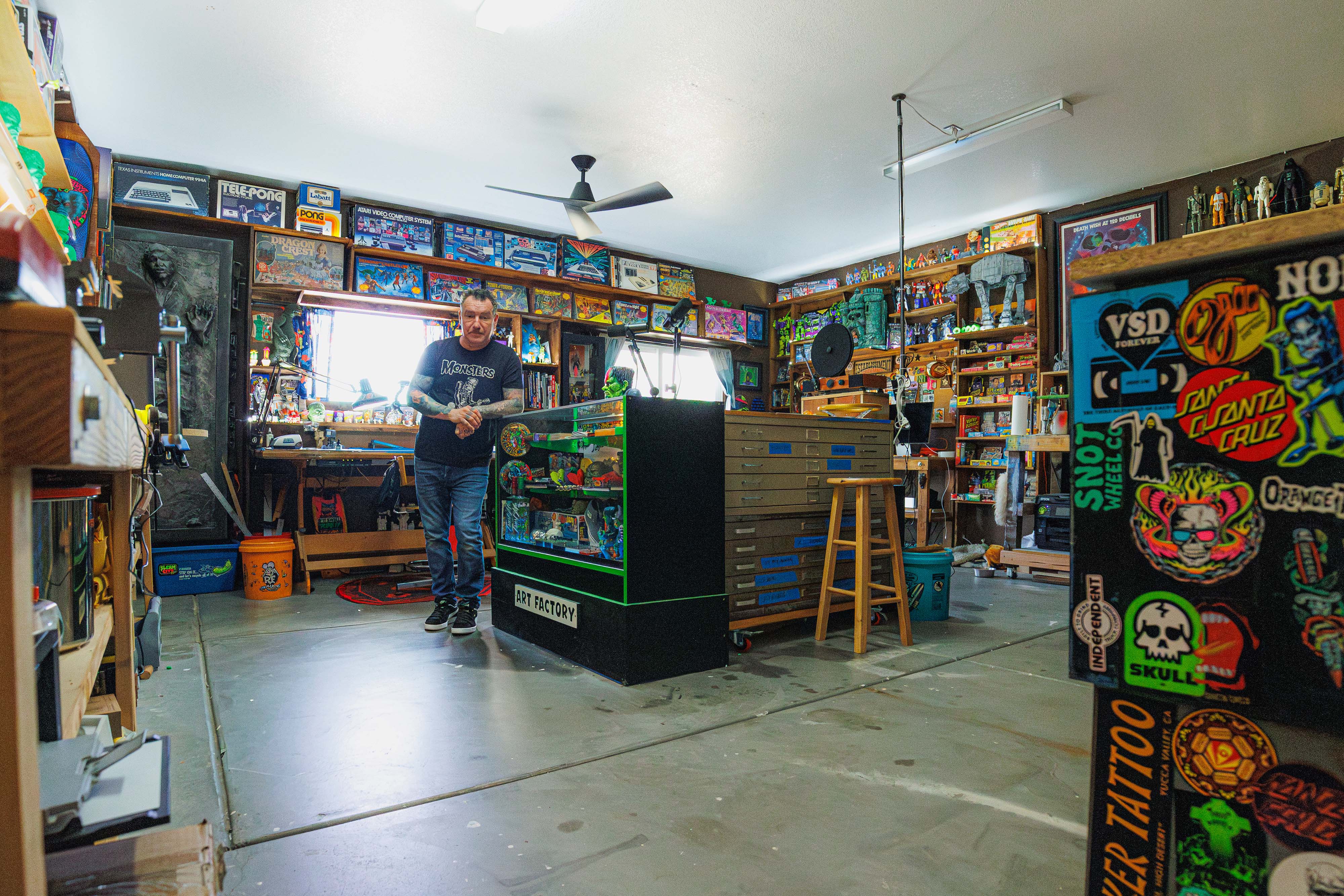 Artist Dirty Donny Gillies standing in his brightly lit studio filled with vintage toys, art supplies, and colorful collectibles, surrounded by shelves packed with models, posters, and pinball machines.