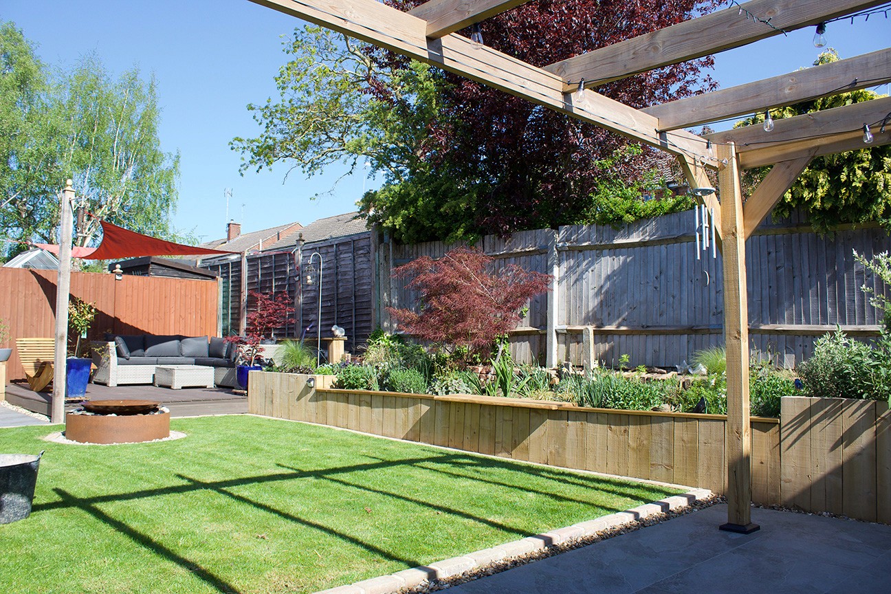 A sunny garden featuring a green lawn, wooden pergola, and landscaped plants against a rustic backdrop.