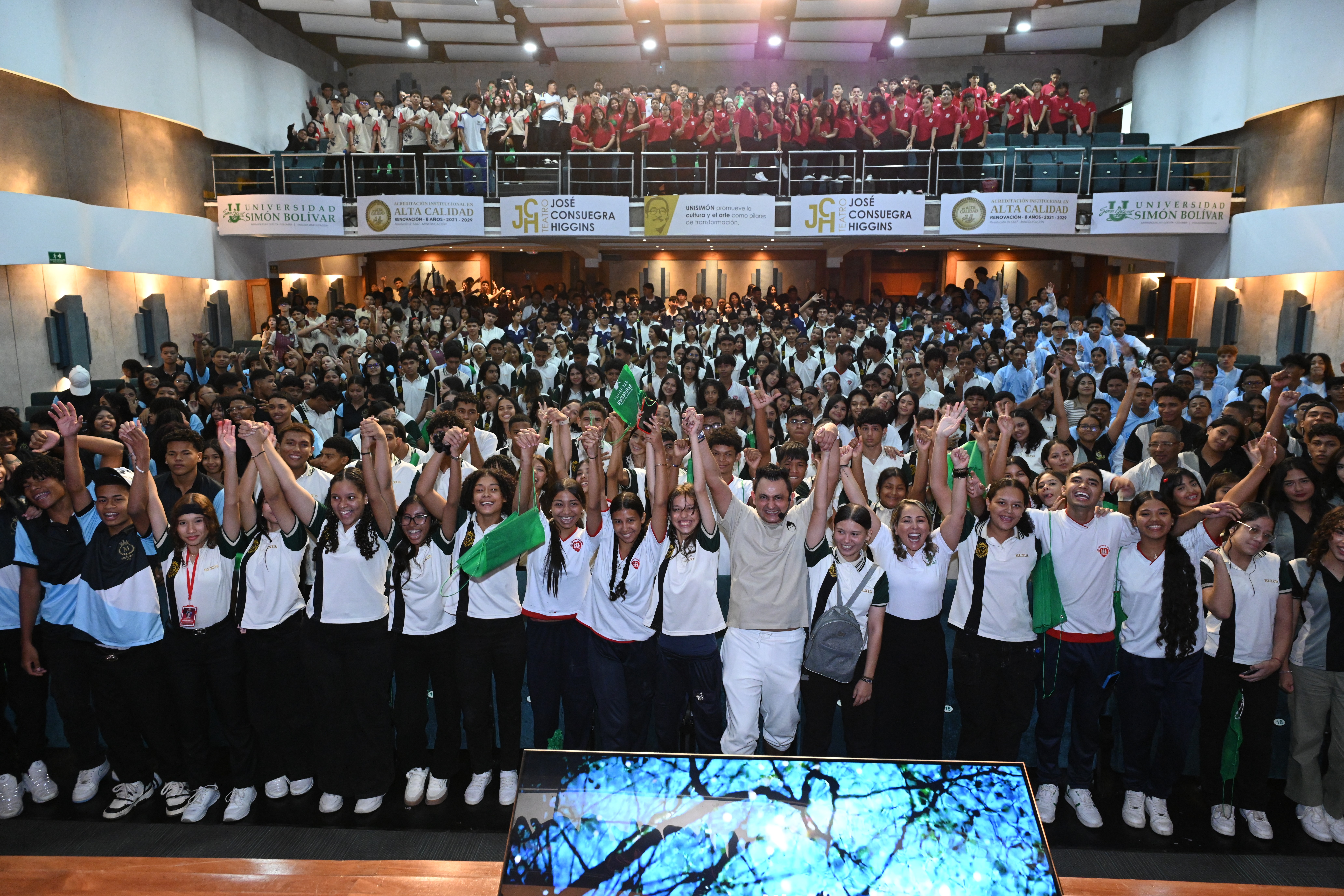 Jorge Baena en el escenario rodeado de estudiantes sonrientes con los brazos levantados, celebrando el cierre de una conferencia motivacional en un auditorio lleno en la Universidad Simón Bolívar.