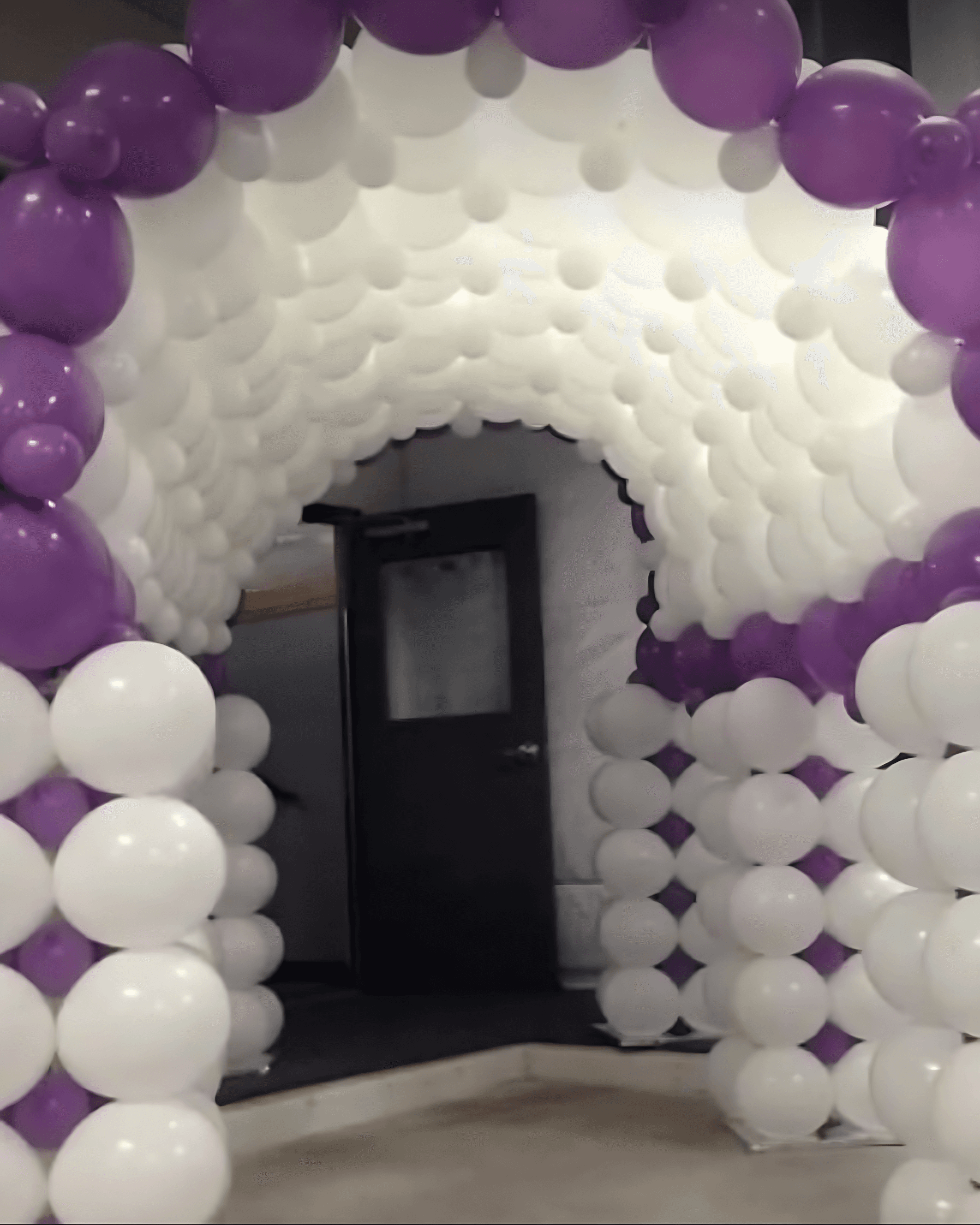 A decorative balloon archway composed of alternating white and purple balloons, leading to a wooden door in a softly lit room.