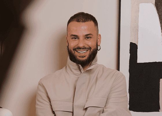 Otávio Lameiro a sorrir para a câmara, de braços cruzados, com barba e brinco de argola, usando um casaco bege