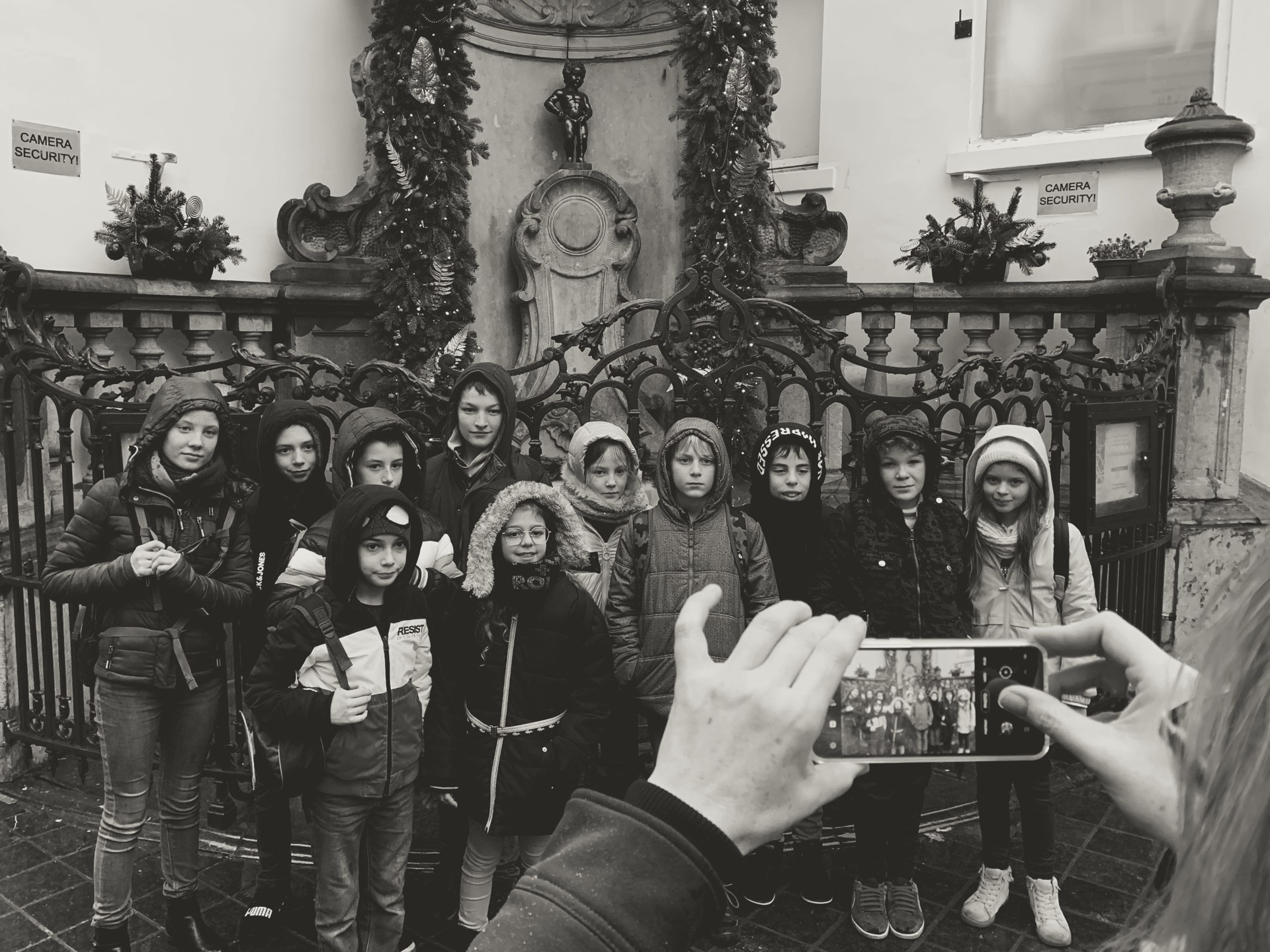 Black-and-white Brussels travel photo of children at Manneken Pis fountain with festive garlands and wrought-iron fence; ideal for content about Brussels landmarks, family travel, and holiday sightseeing.