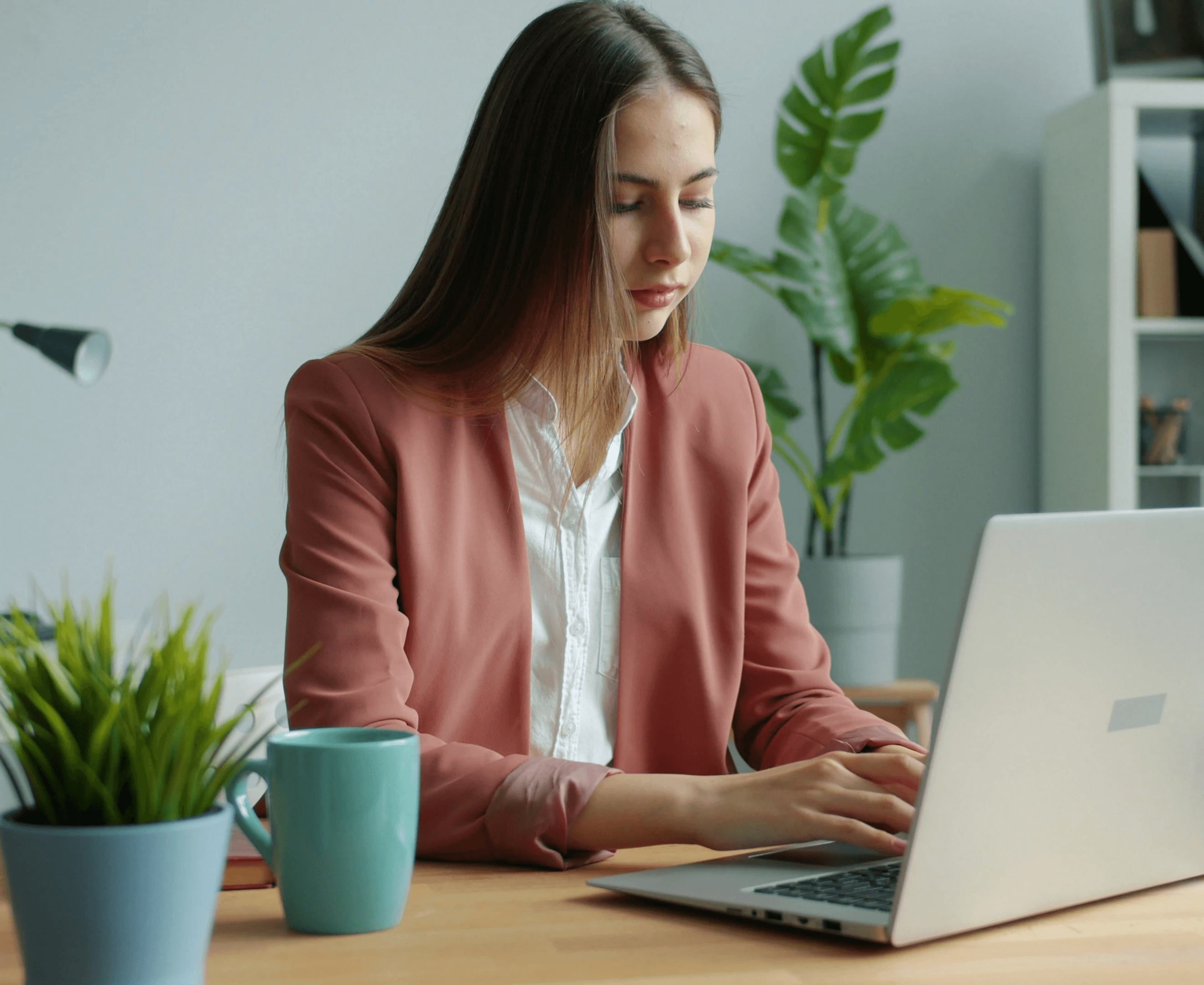 Businesswoman working on laptop at modern office desk with plants and coffee mug
