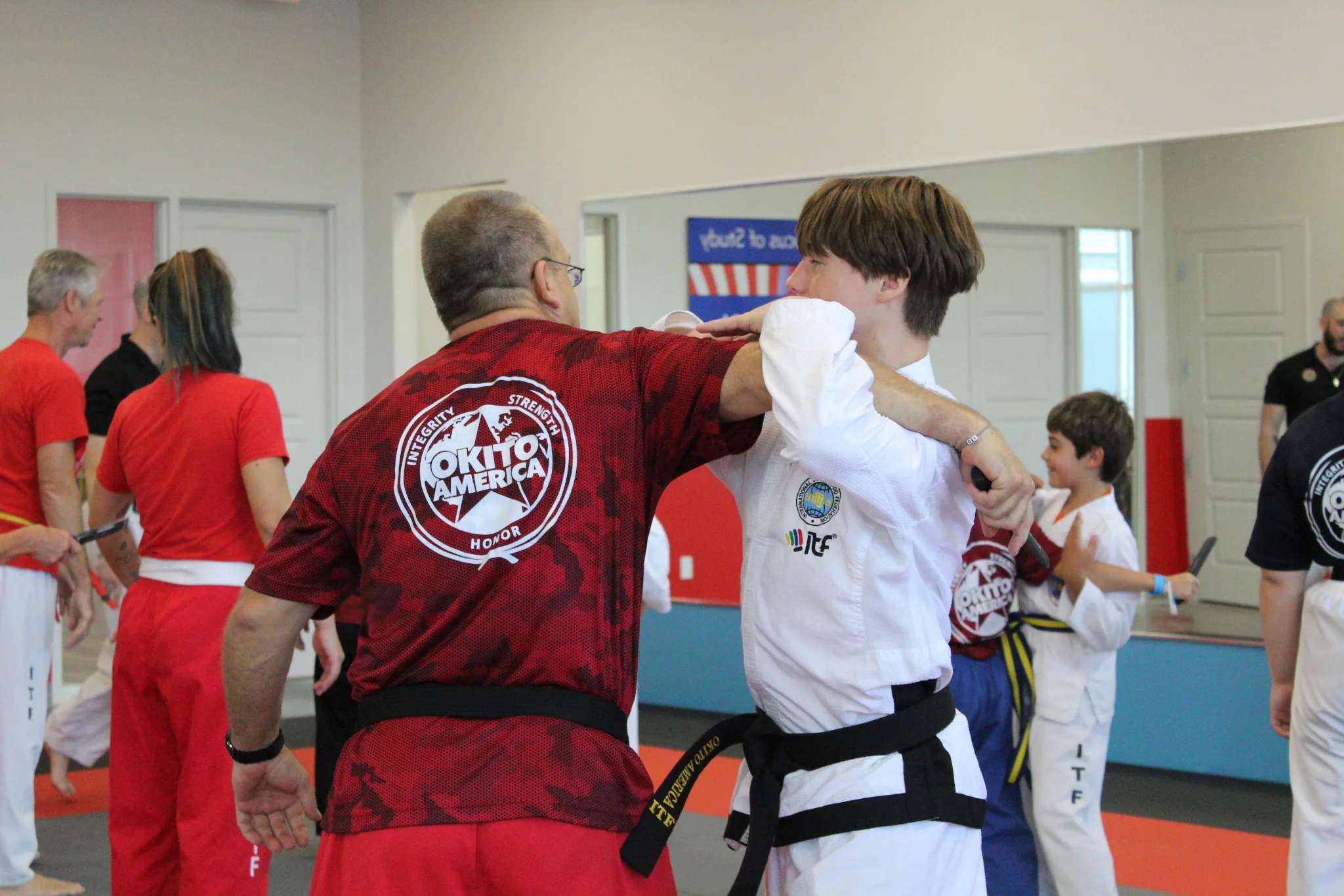 An instructor in a maroon "Okito America" gi demonstrates a martial arts technique by lifting a young student in a white gi during a class session in a dojo, with other students and instructors watching in the background.