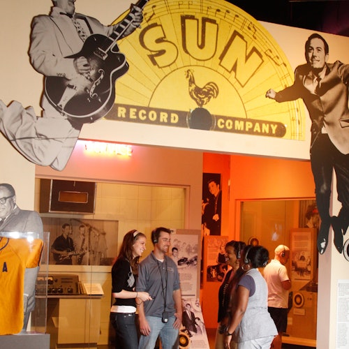 Lidé si prohlížejí exponáty v muzeu pod velkým nápisem „Sun Record Company“, po stranách doplněným fotografiemi hudebníků.