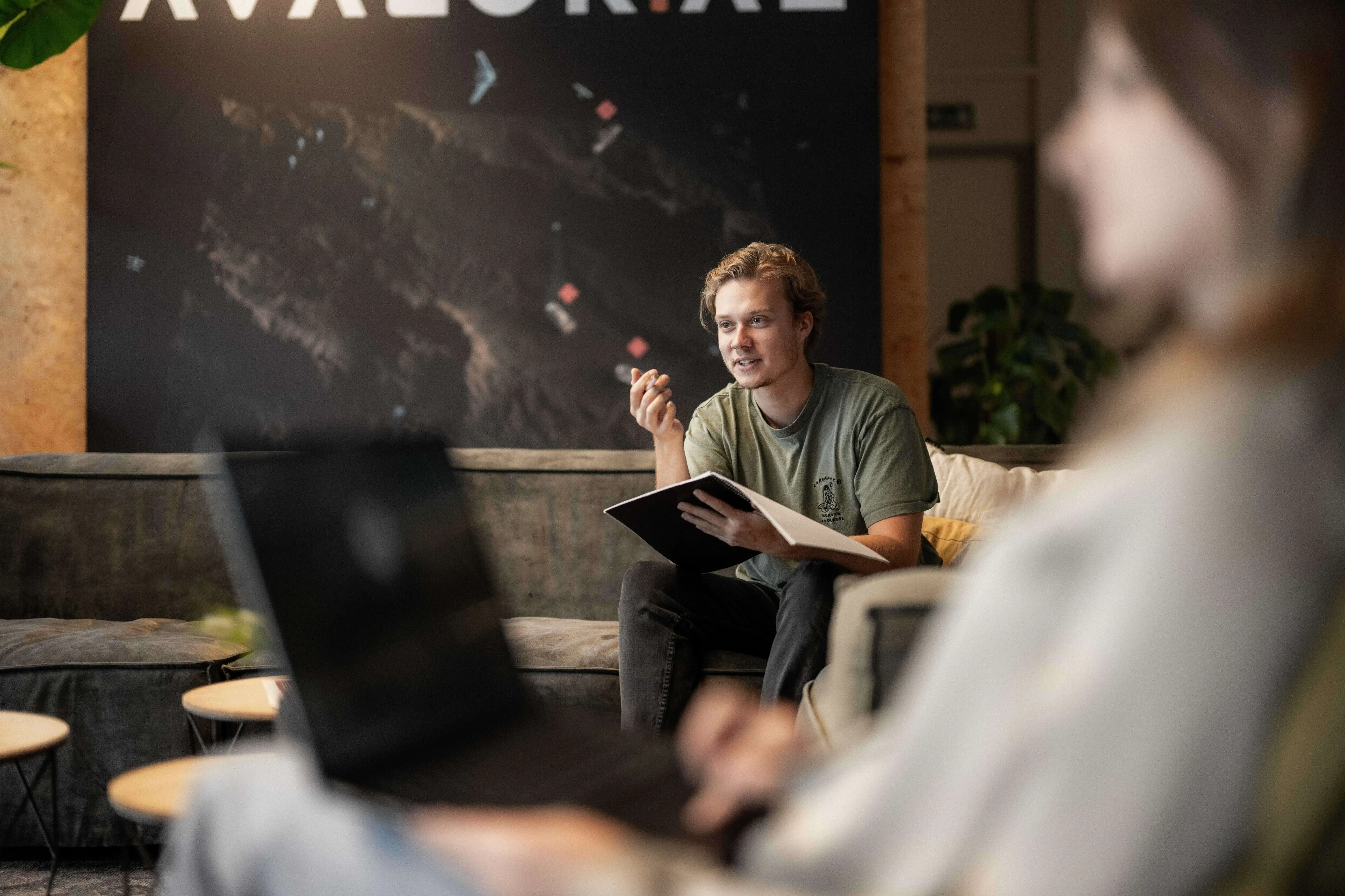 A person with a notebook gestures while seated on a gray couch in a modern, softly lit office space with a large abstract wall art in the background.