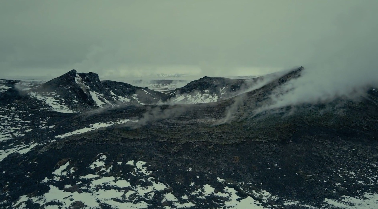 Misty volcanic landscape with jagged, snow-dusted peaks and steaming vents under clouds.