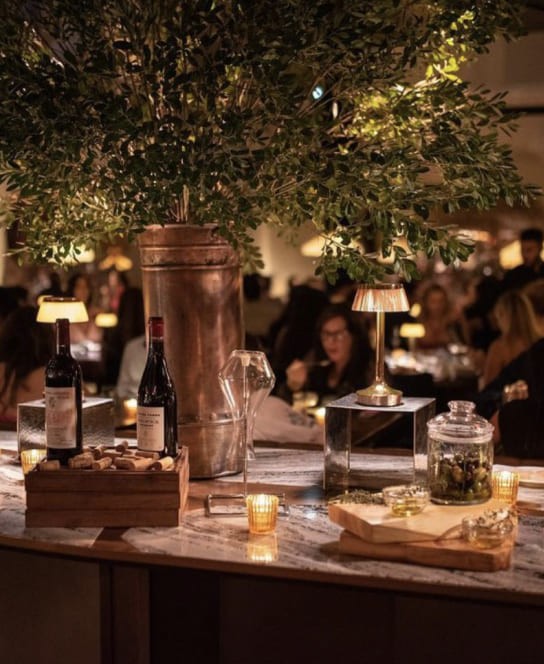 Intimate dining scene with dim lighting, featuring a rustic table set with bottles of wine, a copper pitcher, and small lamps amidst a lively restaurant backdrop.