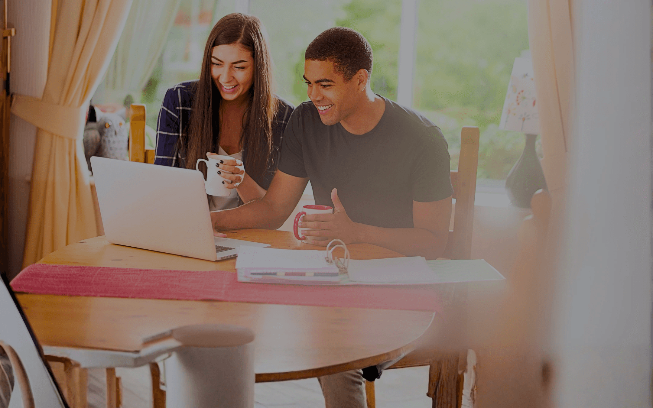 Couple smiling while looking at a laptop computer together at a round wooden table, holding mugs and documents.