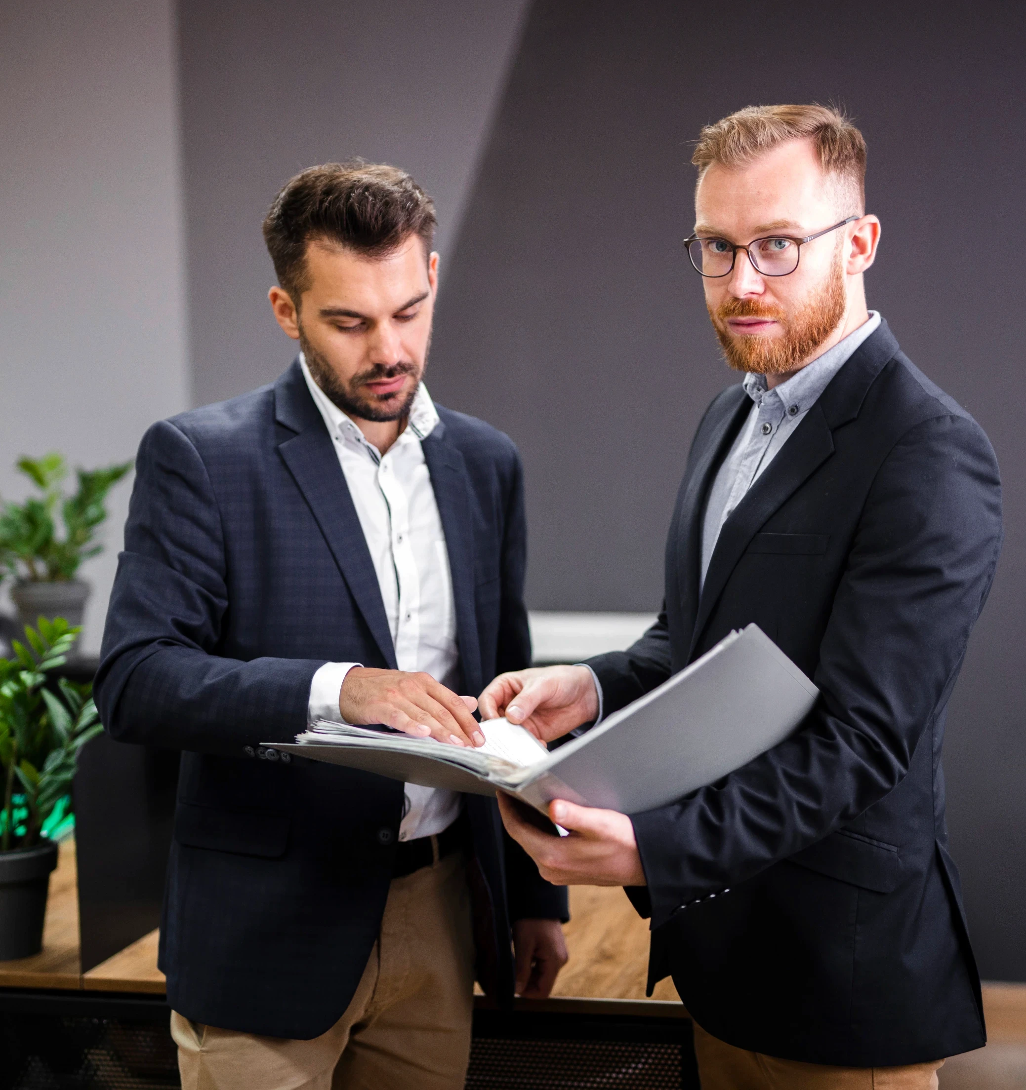 Two men in suits reviewing documents in a modern office setting. One points at a page, while the other holds the folder, focused and professional.