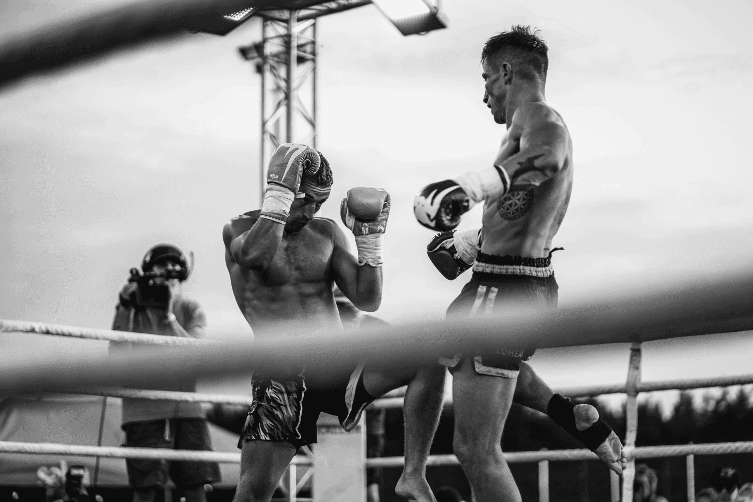 Two shirtless fighters exchanging a kick during a kickboxing or Muay Thai match inside a boxing ring, with gloves on and a cameraman filming in the background.