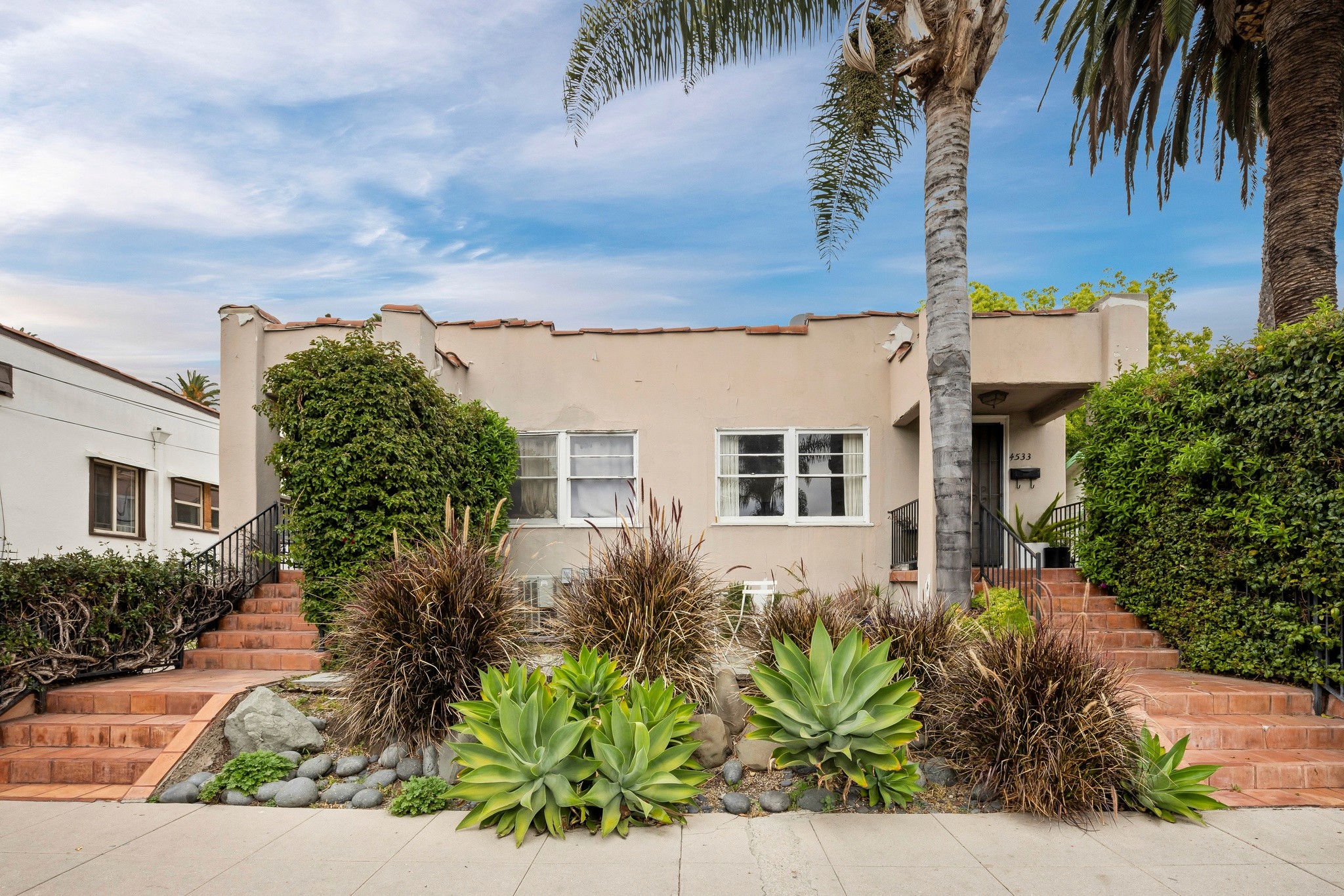 A light-colored house at 4533 ambrose ave, surrounded by lush greenery, palm trees, and a clear blue sky.