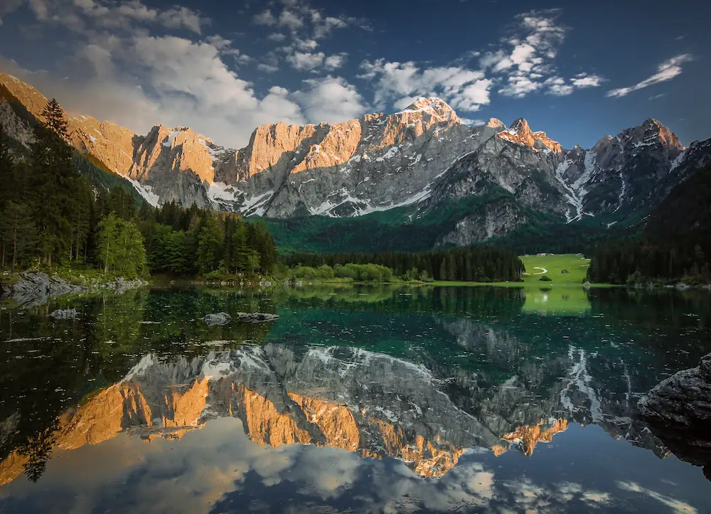 A perfect mirror reflection of the rugged, sunlit Mangart mountains in the calm waters of Lago di Fusine, Italy, surrounded by dense evergreen forests.