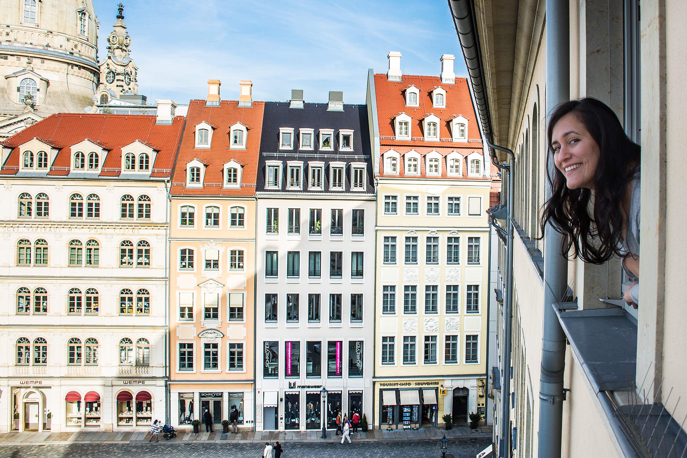 A woman leans out of a balcony window smiling at a row of colorful historic buildings in a European city square.