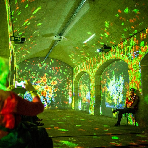 Pessoas observando projeções digitais coloridas nas paredes e no teto de um túnel arqueado, com uma pessoa sentada em uma cadeira.