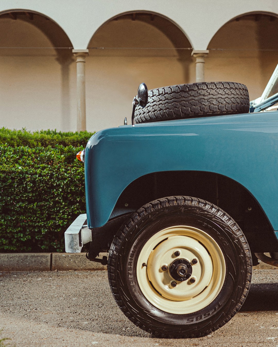 Side detail of a vintage blue Land Rover Series II, showing the front wing, cream-painted steel wheel and a spare tyre laid on the bonnet, parked on gravel before an arched colonnade and a hedge.