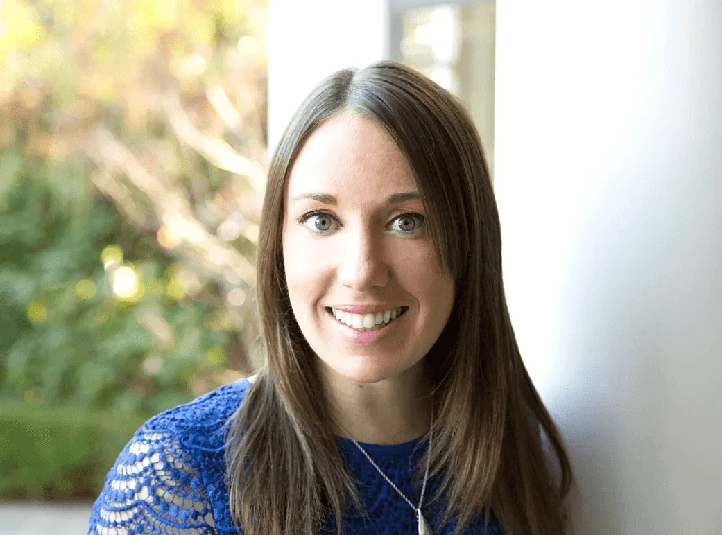 Professional woman with long brown hair smiling confidently, wearing a blue lace top and silver necklace.