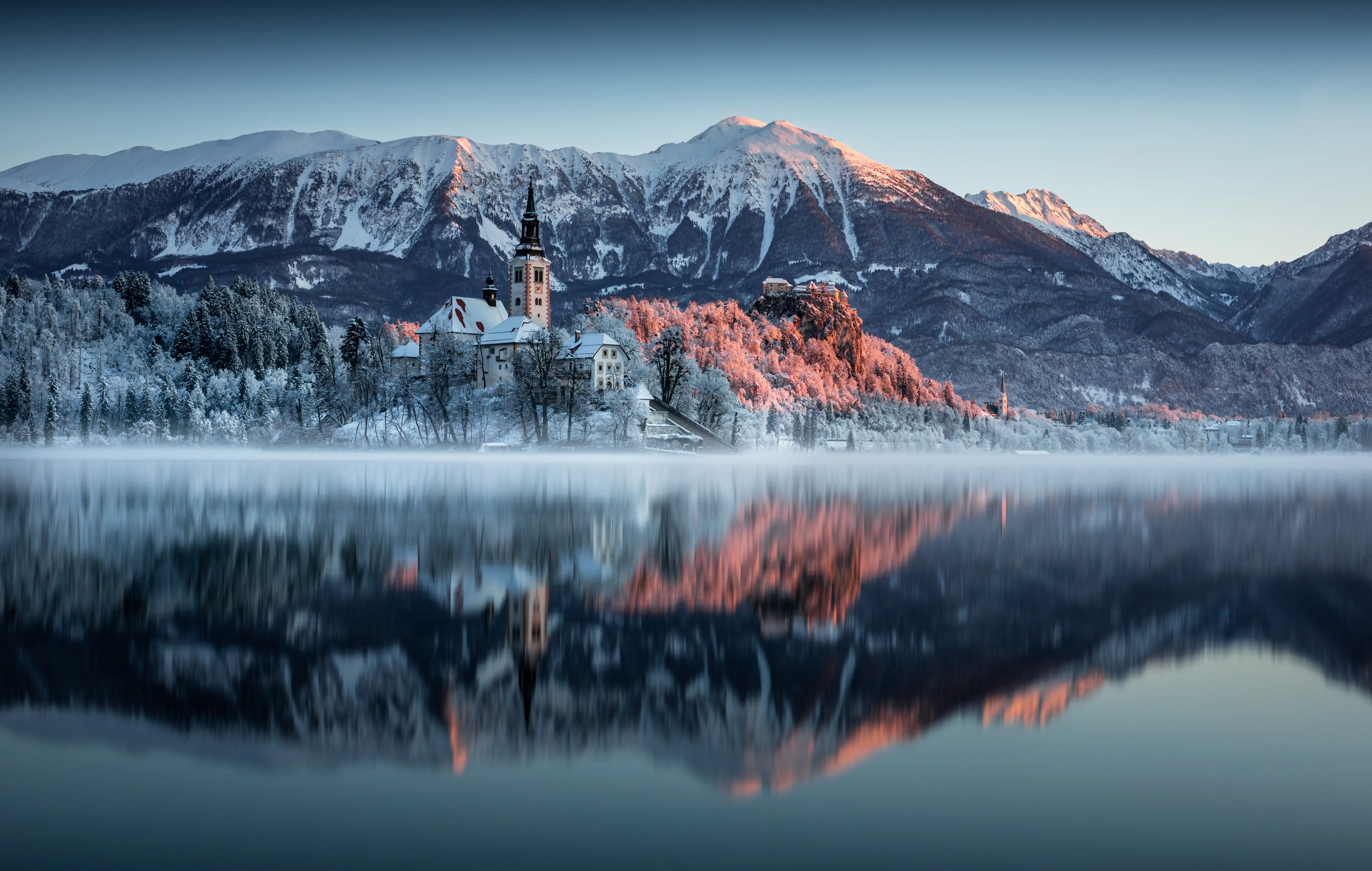 Lake Bled in winter, featuring the island church and clifftop castle reflected in frozen-looking, calm water under sunlit, snow-capped mountain peaks.