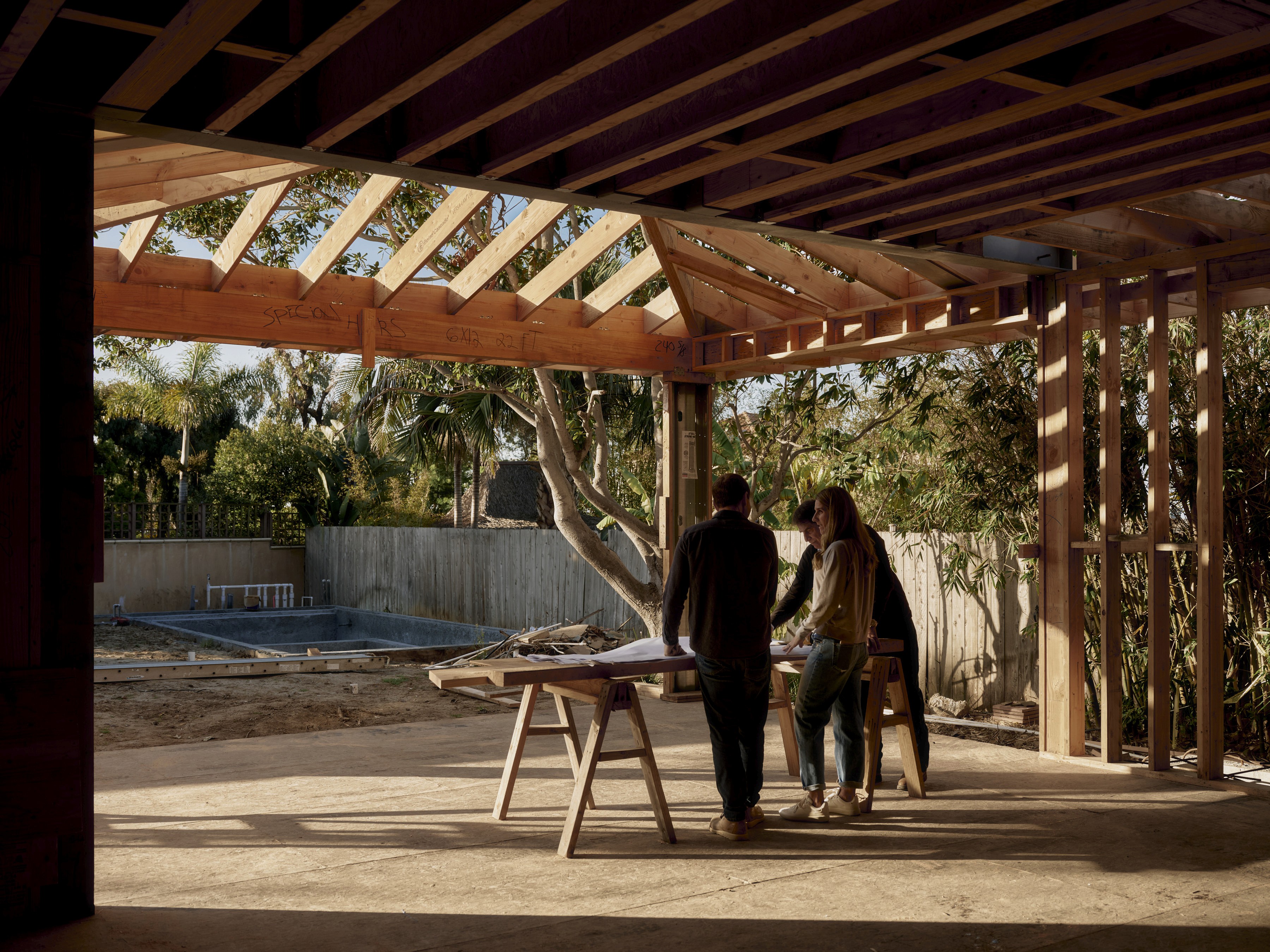A group of professionals reviews blueprints on a job site within a residential wood-framed structure. An outdoor construction area with a pool shell is in the background.