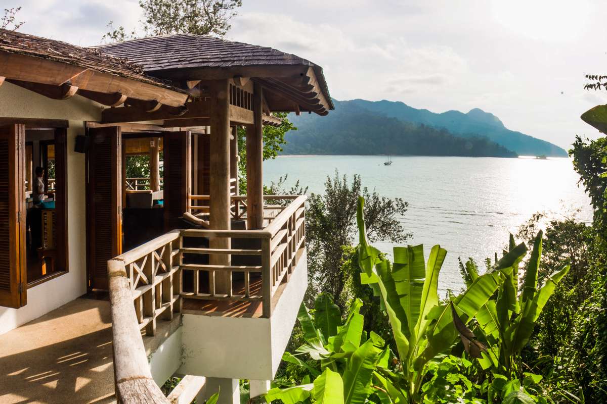 A wooden beach bungalow with a thatched roof overlooks a turquoise ocean and green mountains under a bright sky.
