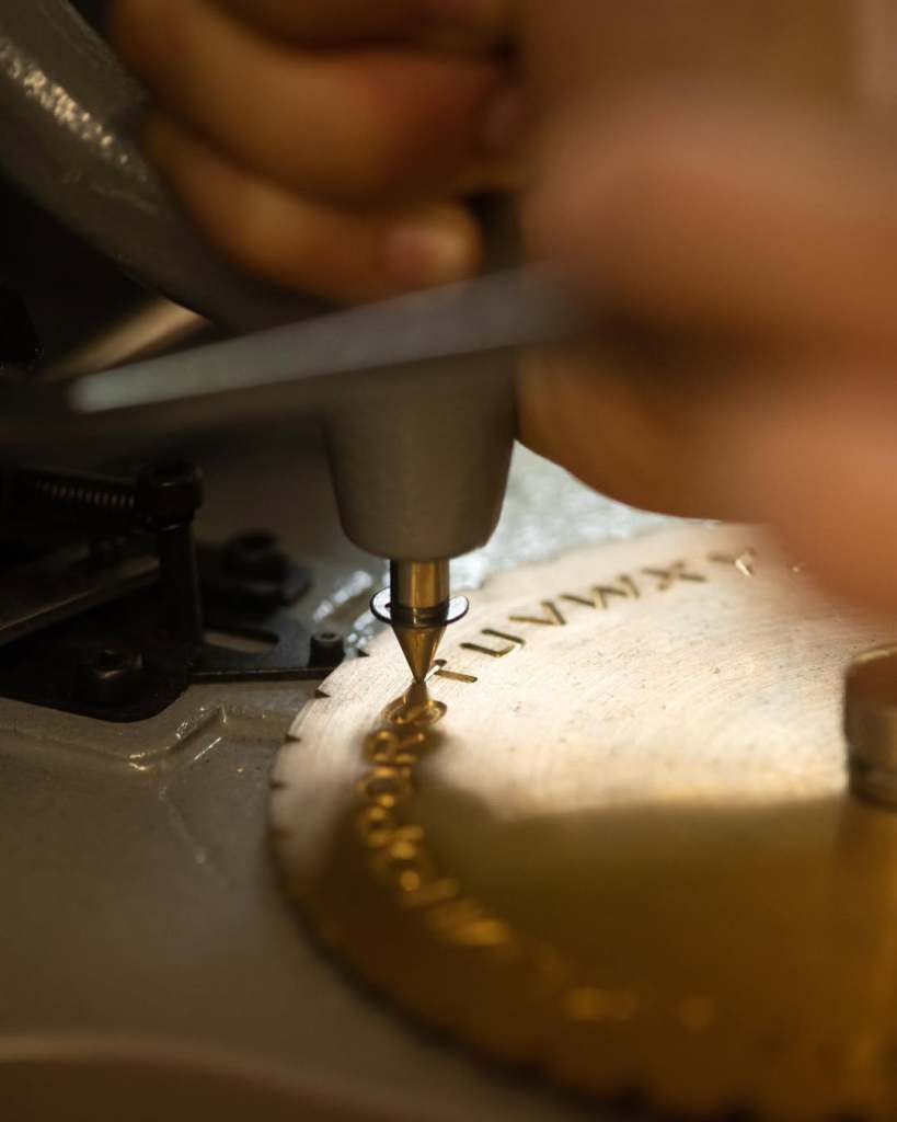 A close-up of a hand operating a engraving tool on a metal disc, showing intricate lettering being created.