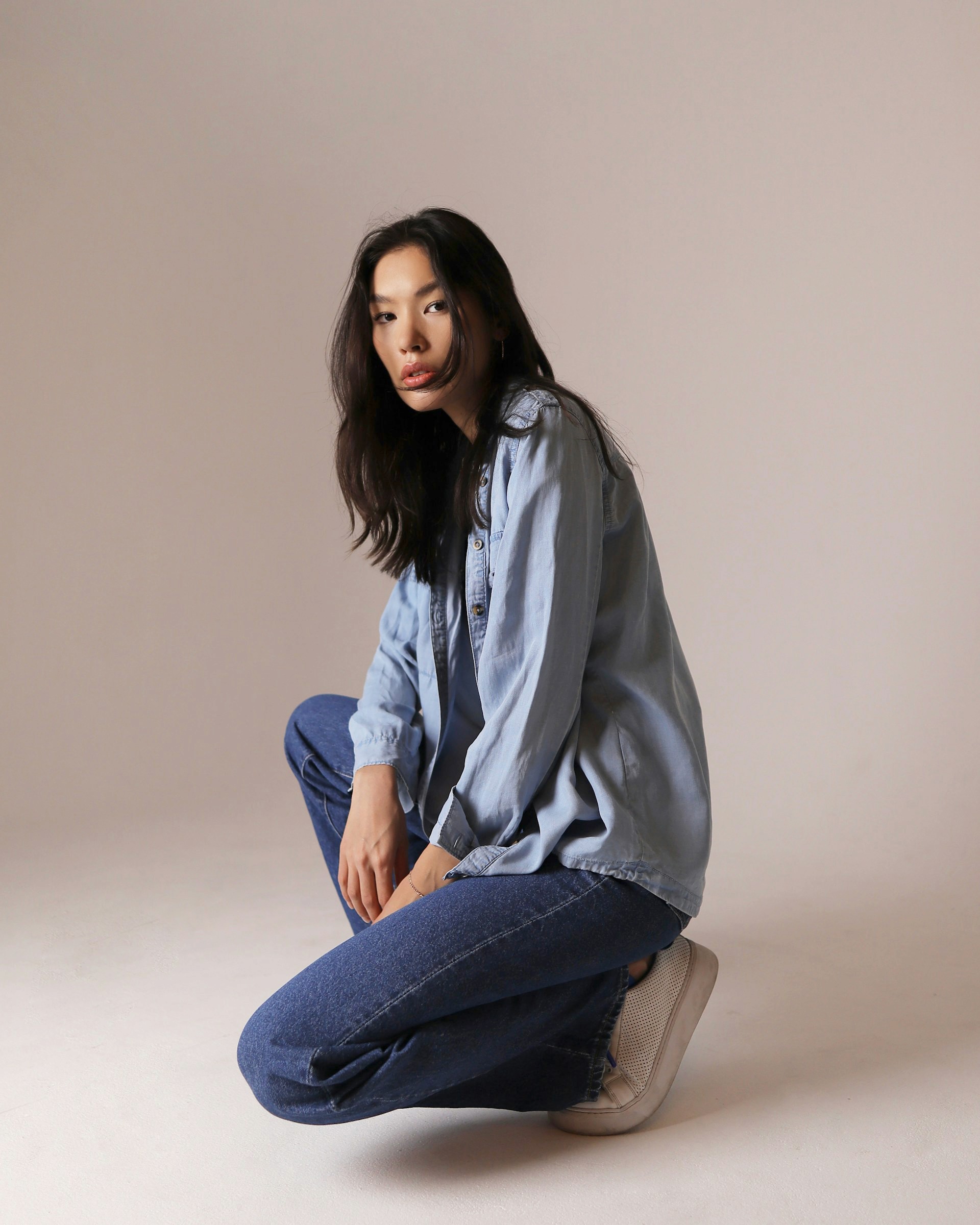 A young woman with long dark hair crouching in a light blue button-down shirt and dark blue jeans against a neutral studio background.