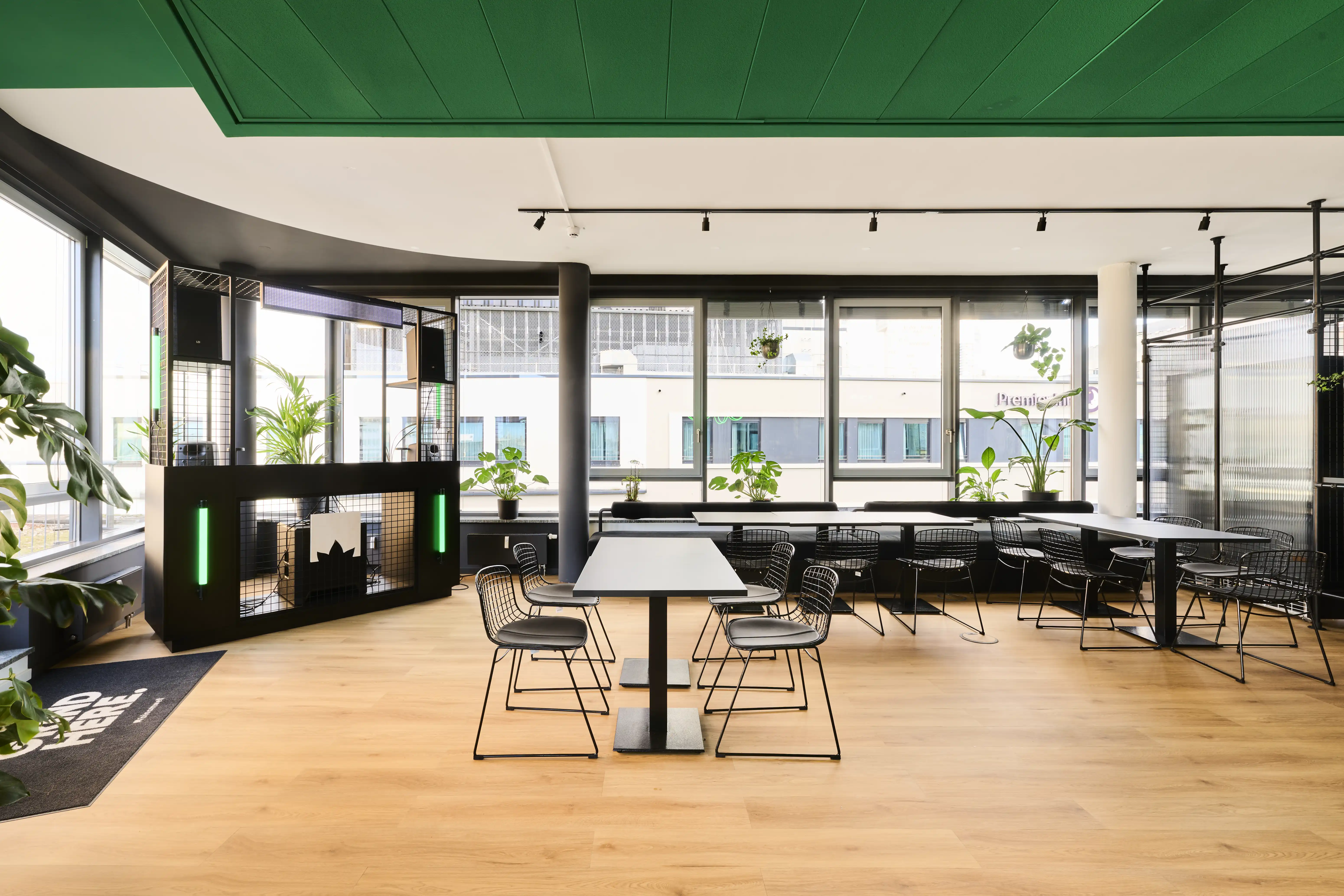Work café at BLOOMWELL with an emerald-green ceiling, black wire chairs and bistro tables, window benches with black cushions, plants and the black mesh DJ booth.