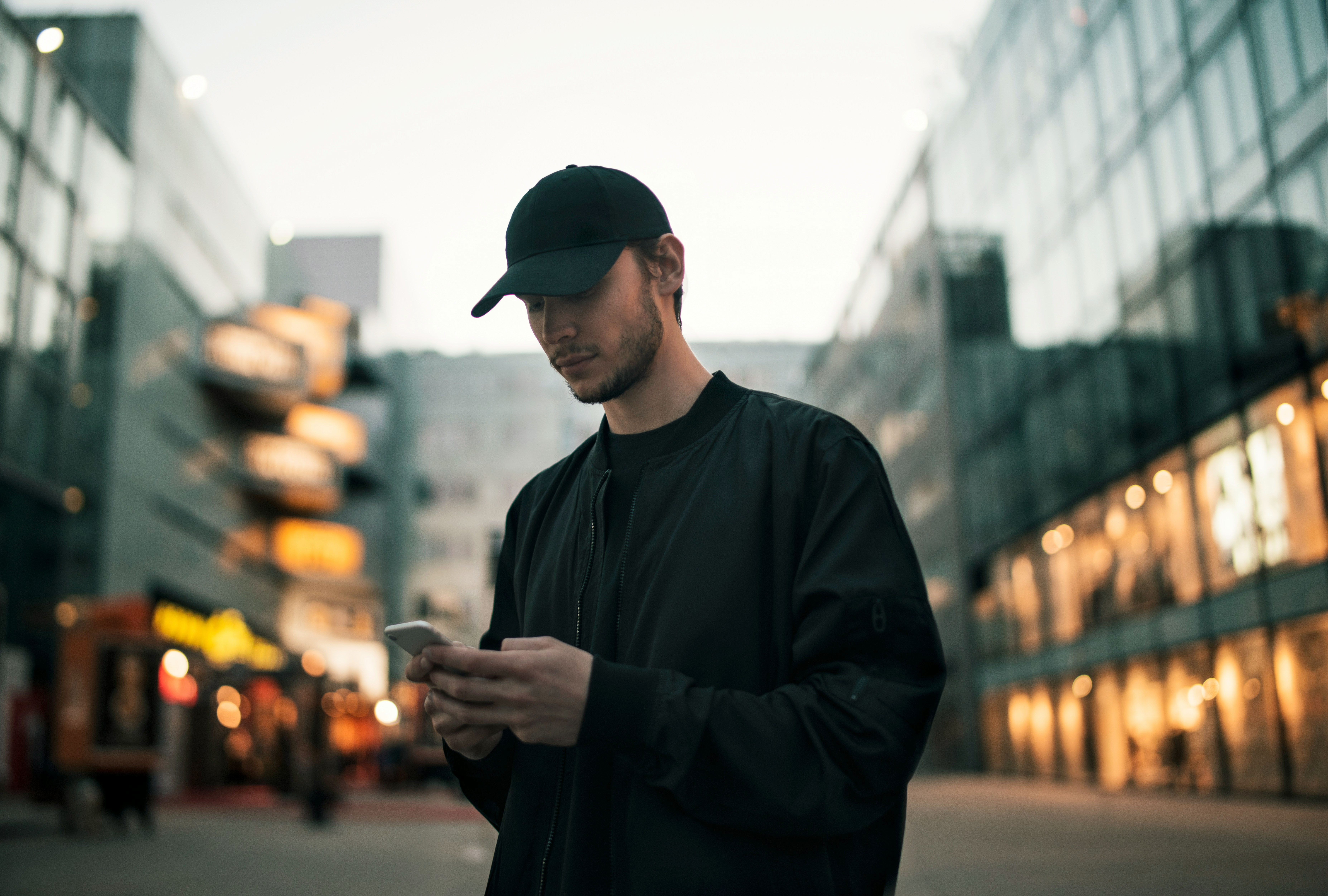 A person in a black outfit and cap stands outdoors, holding a phone, surrounded by modern buildings.