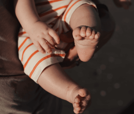  A close-up of a person's hands holding a small child, whose feet are dangling. The child is wearing a striped outfit, and the focus is on their bare feet, which are slightly sandy. The background is blurred, suggesting an outdoor setting, possibly at the beach.