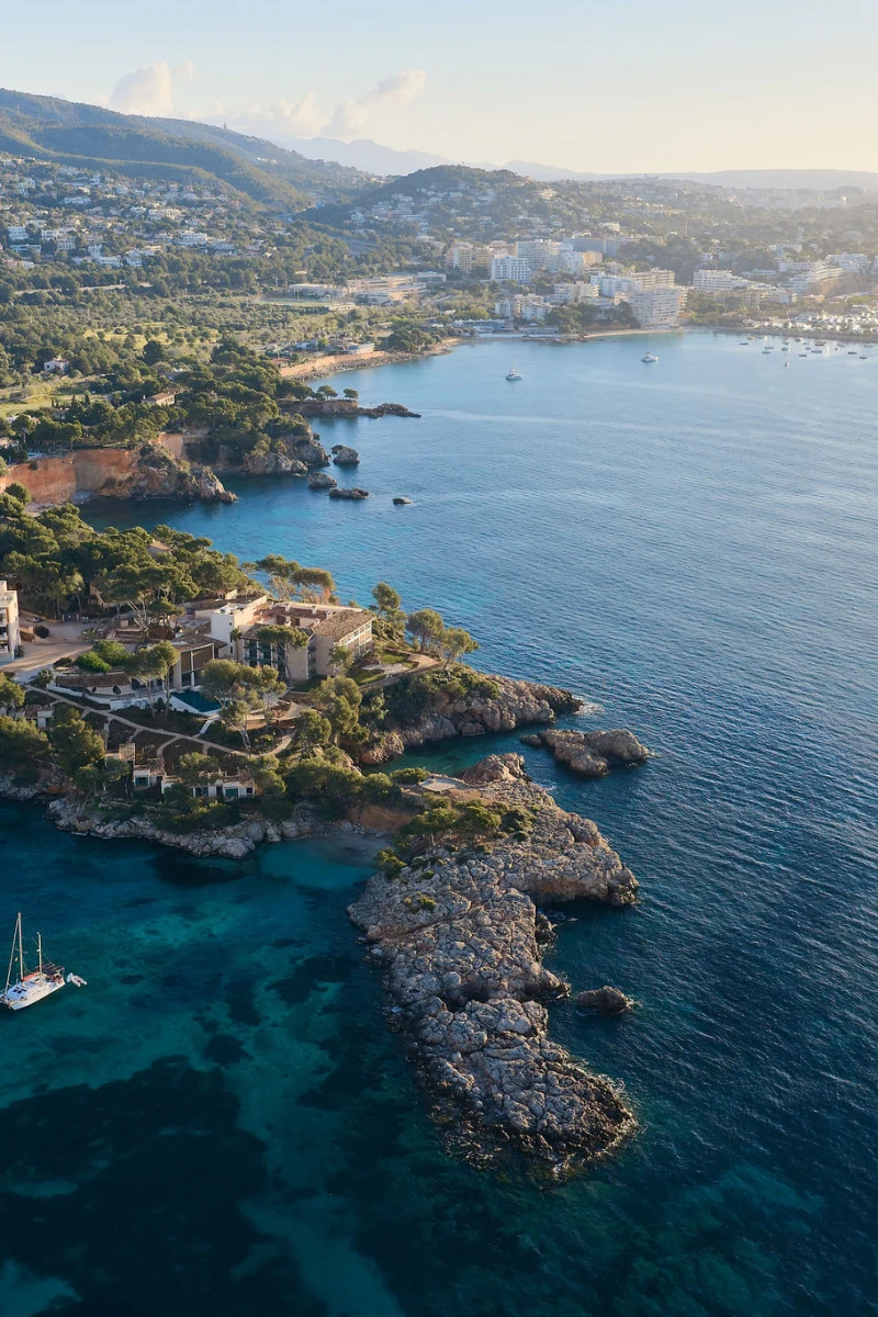 in Blick aus der Luft auf ein luxuriöses Küstenresort mit einem großen Pool, das auf einer felsigen Halbinsel liegt, mit einer Stadt und Bergen im Hintergrund.