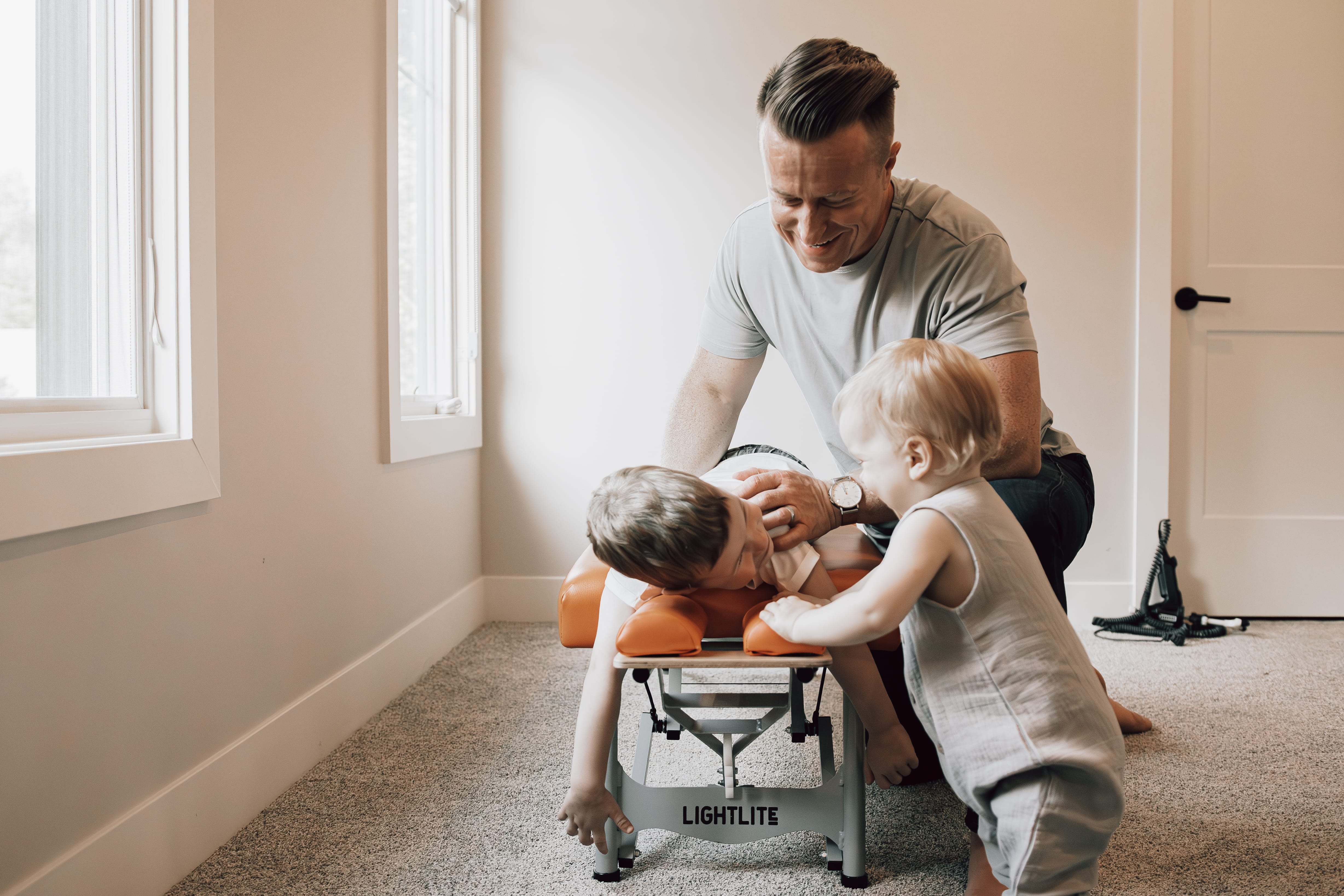 A man smiling and kneeling on the floor playing with laughing children inside a home.