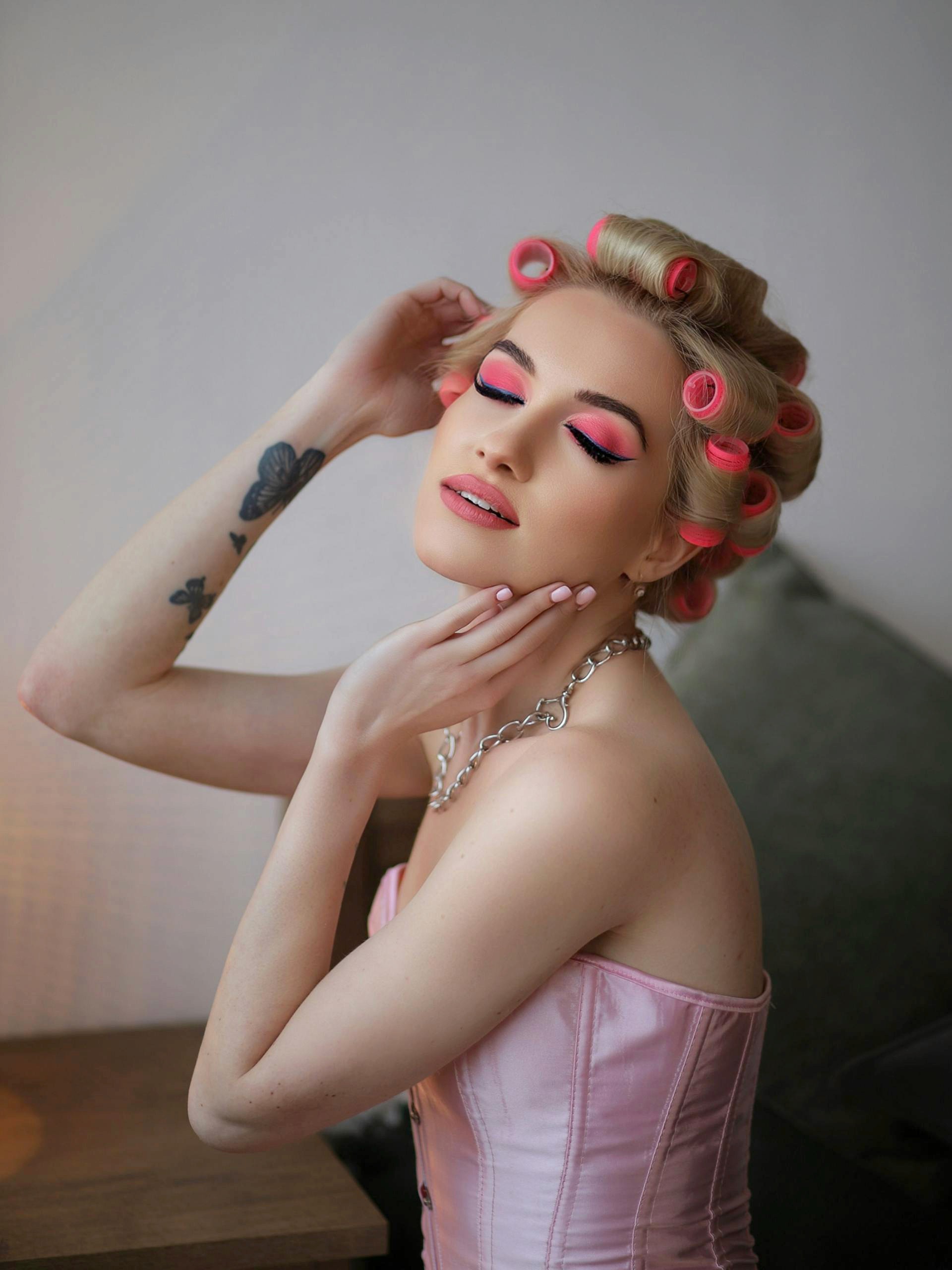 A woman with blonde hair in pink rollers and a pink corset posing with her eyes closed, showing a butterfly tattoo on her arm.