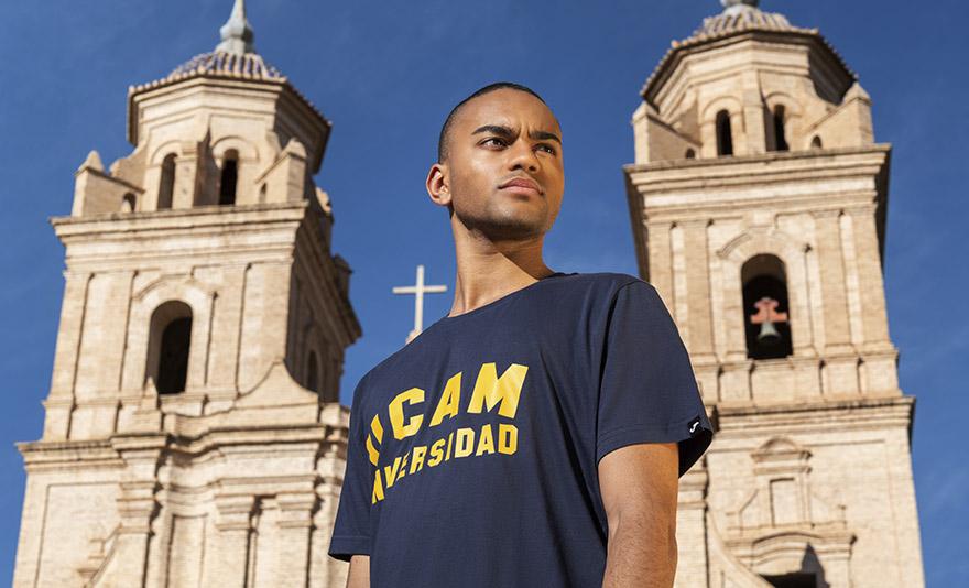 A person stands confidently in front of an ornate, historic church with twin bell towers under a clear blue sky, wearing a navy shirt displaying the letters "UCAM Universidad."