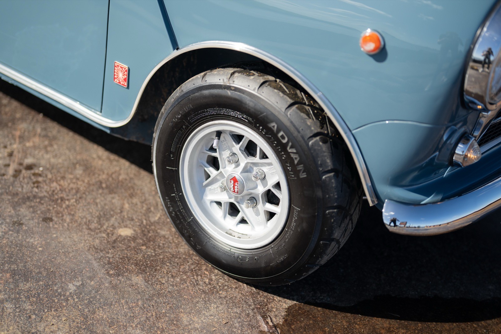 Close-up of a blue car's wheel and tire on a sunny pavement, focusing on the white alloy rim and tire tread.